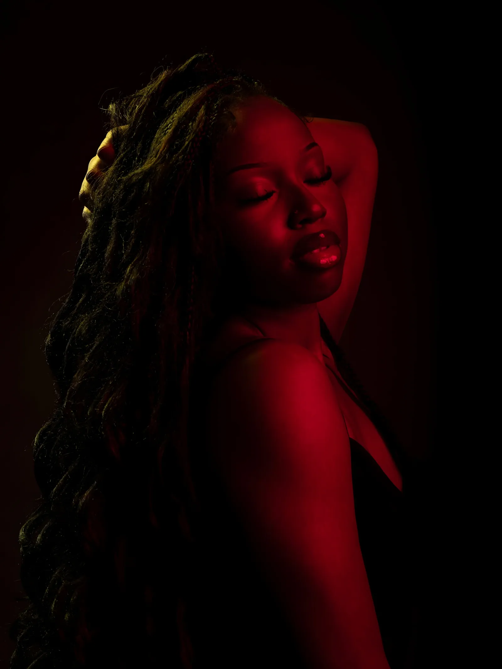 Woman with long braids, eyes closed and one arm raised behind her head, lit by red gel light on black