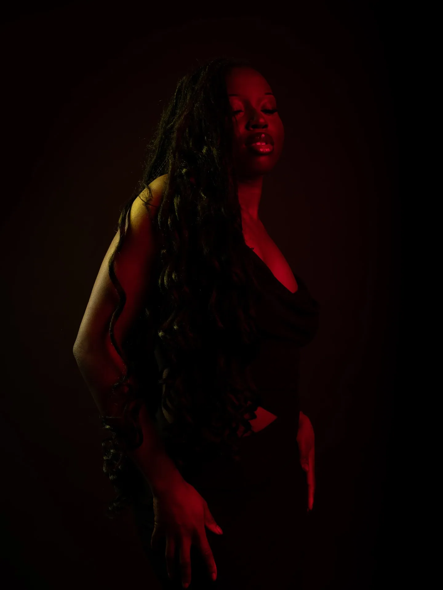 Woman with long wavy hair standing in dark red light, one hand lowered, dark background