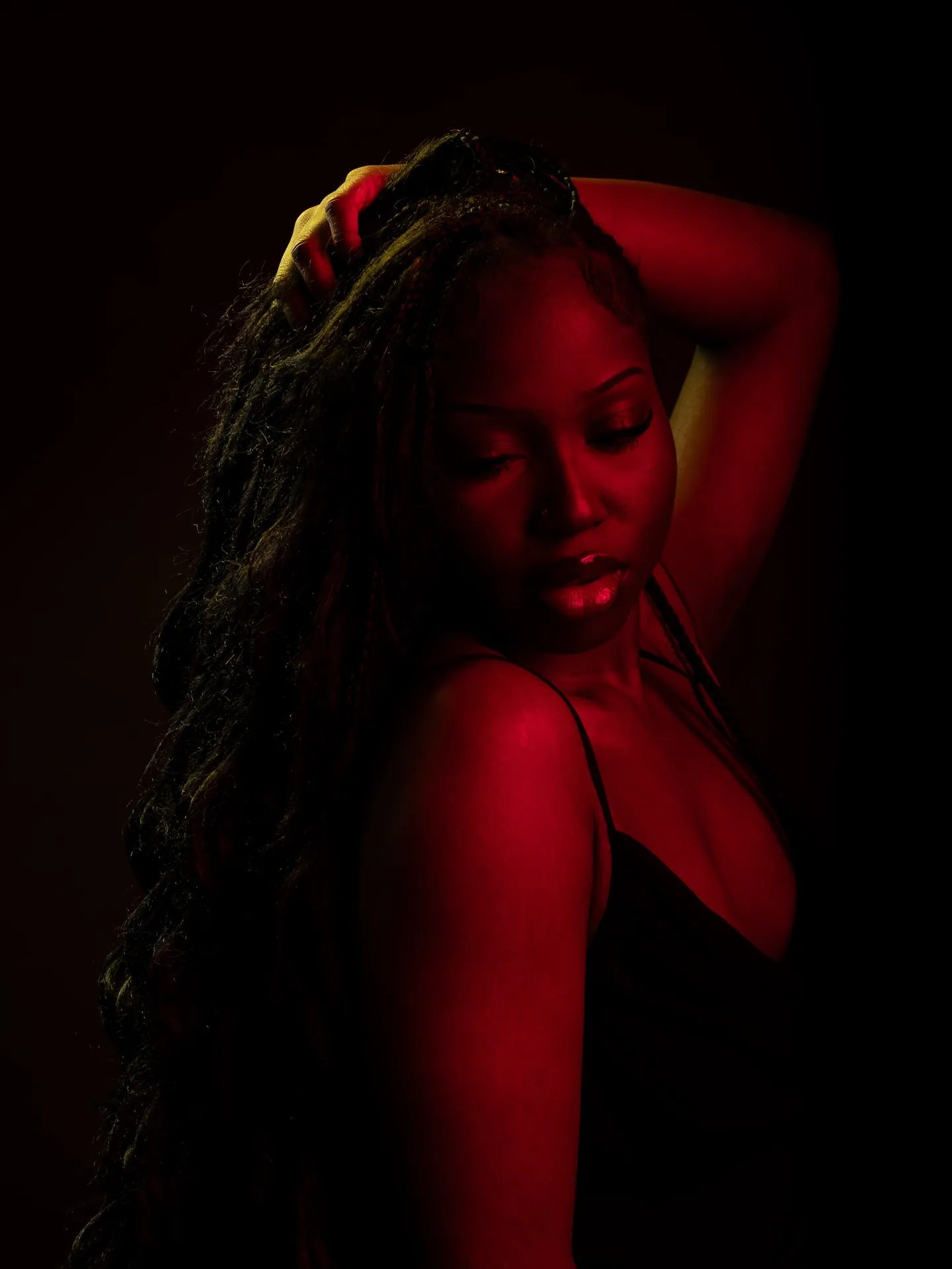 Woman in a black strappy top looking down with a hand in her hair, lit by red gel light