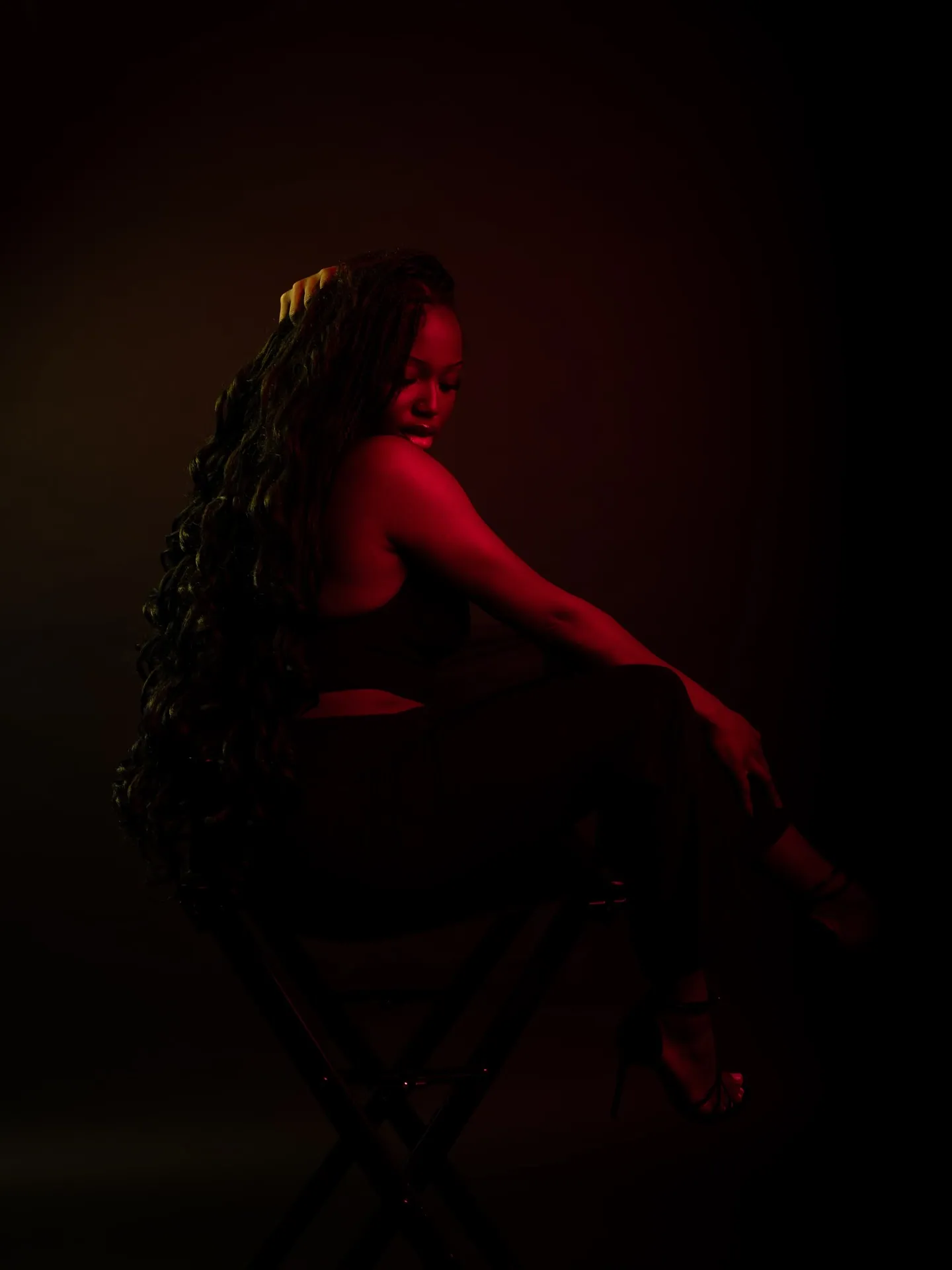 Woman with long hair seated on a stool, glancing over her shoulder, a dim red glow on a dark backdrop