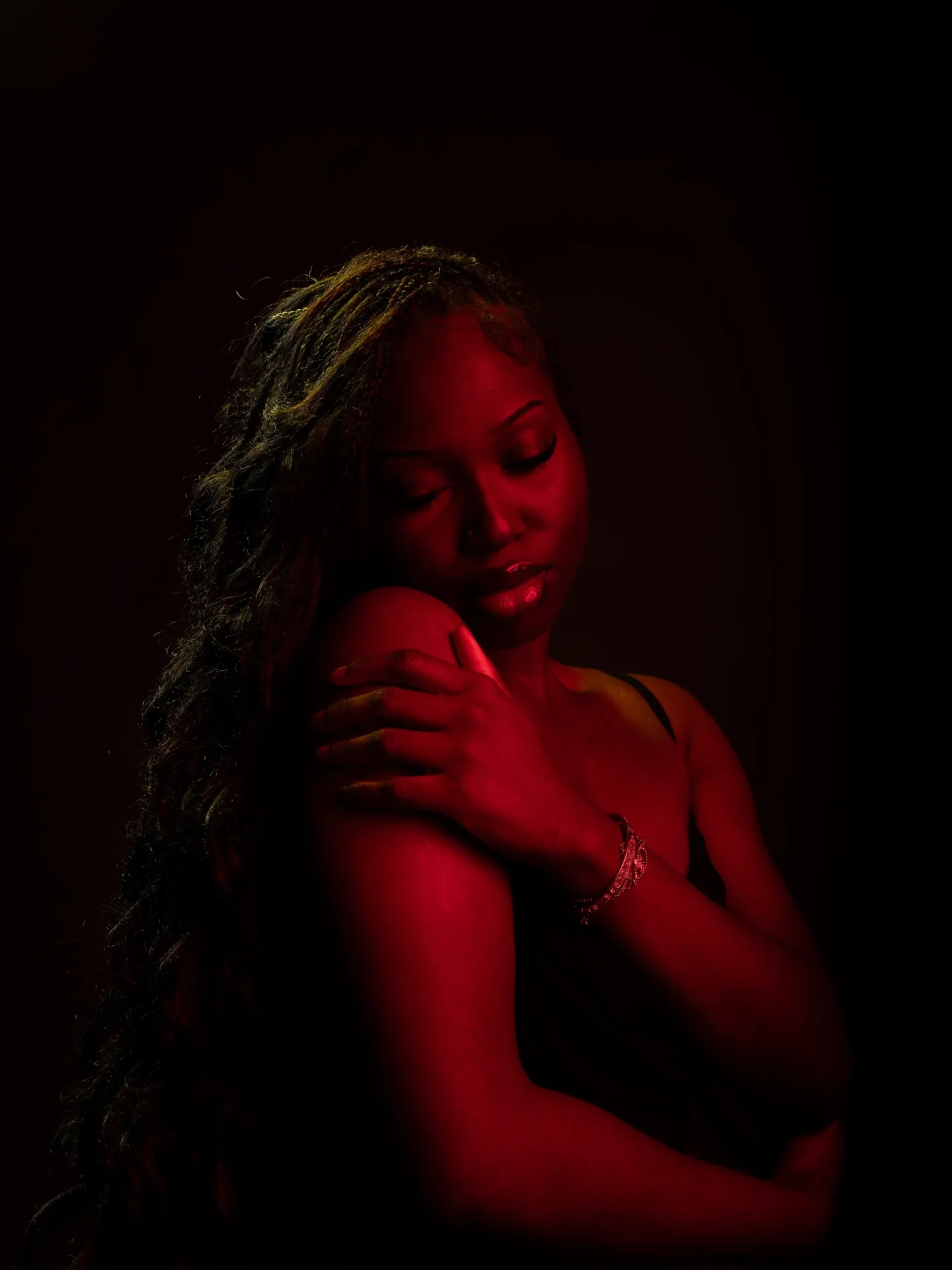 Woman hugging her own shoulder with a bracelet on her wrist, head tilted down, in deep red light