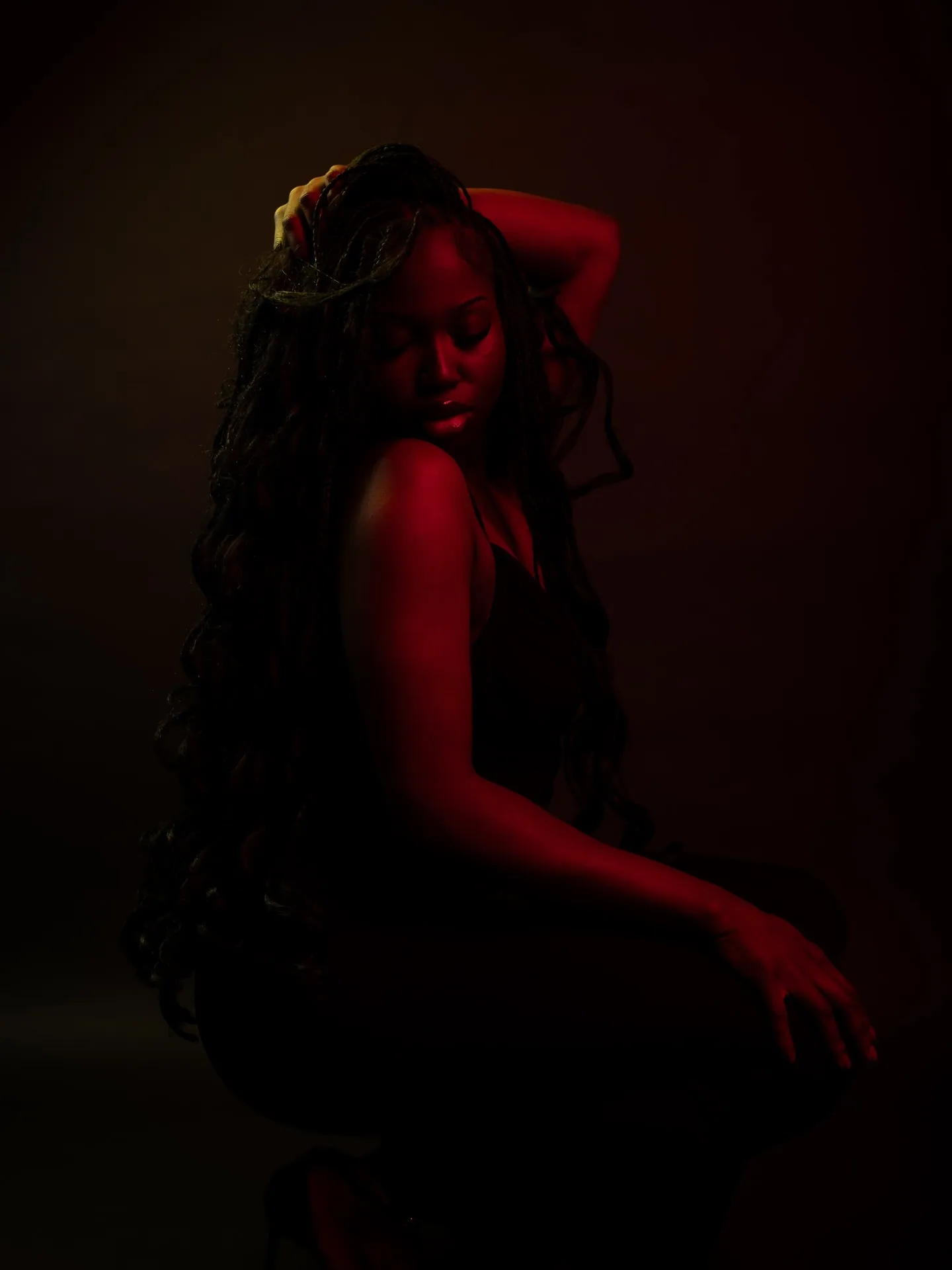 Woman with long hair posed side-on with an arm over her head, in dim red light on black