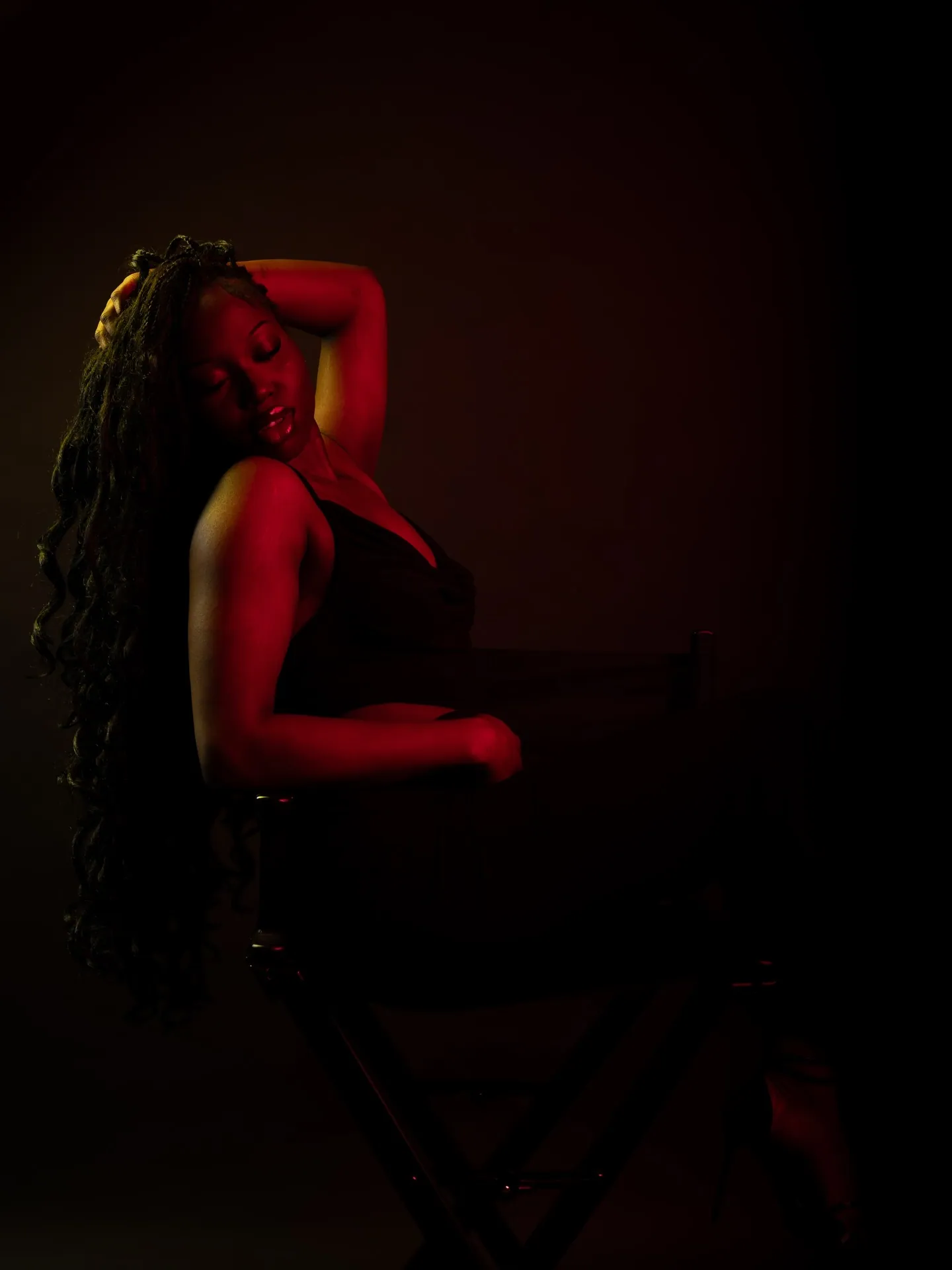 Woman seated on a director chair, arm raised over her head, in red light with a black backdrop