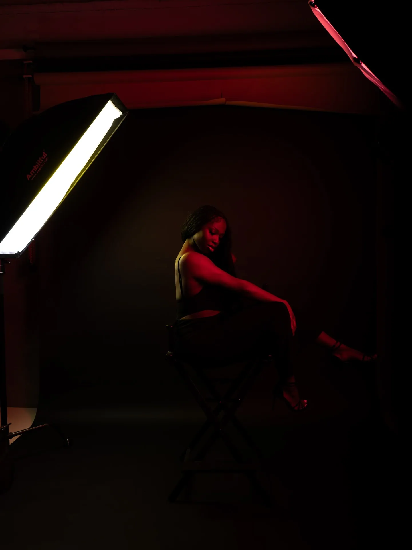 Behind the scenes: woman seated on a stool in a dark red-lit studio, a lit strip softbox at the left