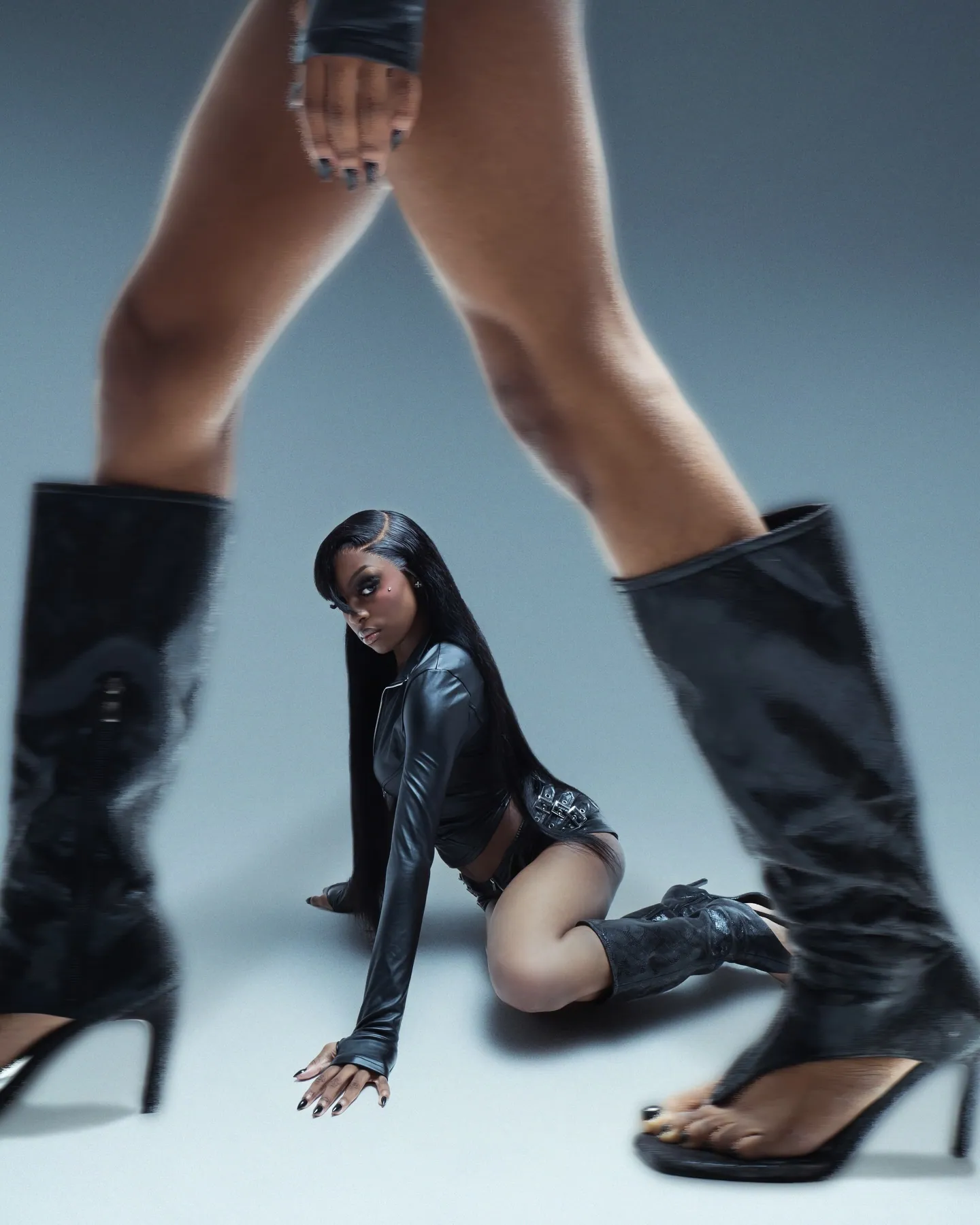A woman in black leather crouching on the floor between the blurred legs and tall black boots of a figure towering over her