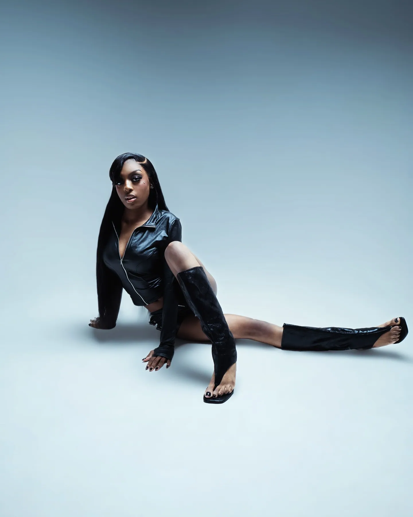 A woman in a black leather jacket and thigh boots sitting back on one arm with legs stretched out, grey-blue backdrop