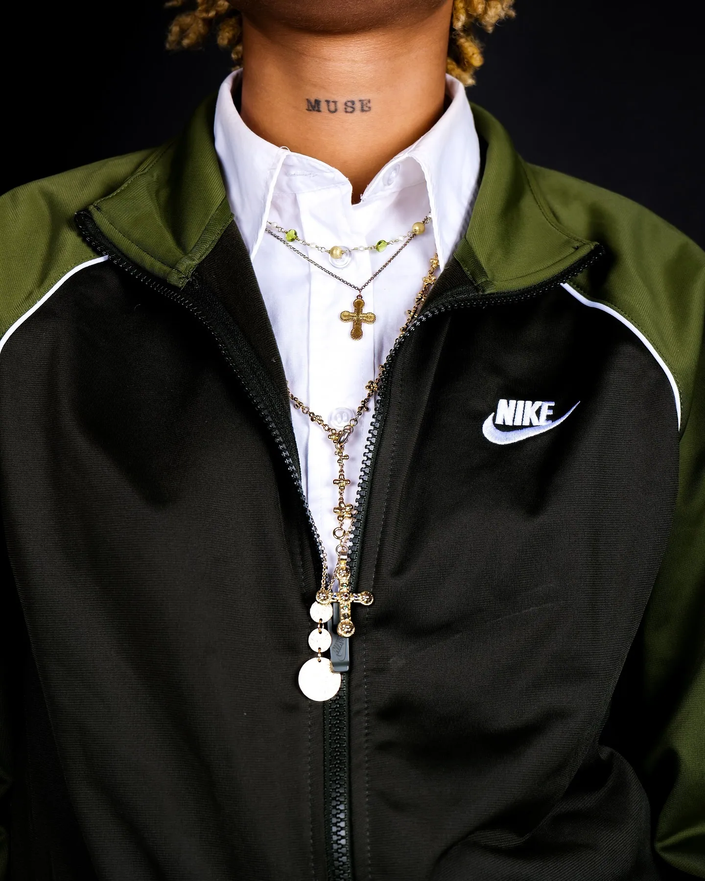 Close crop of a green and black Nike jacket over a white shirt with gold cross necklaces and a MUSE neck tattoo