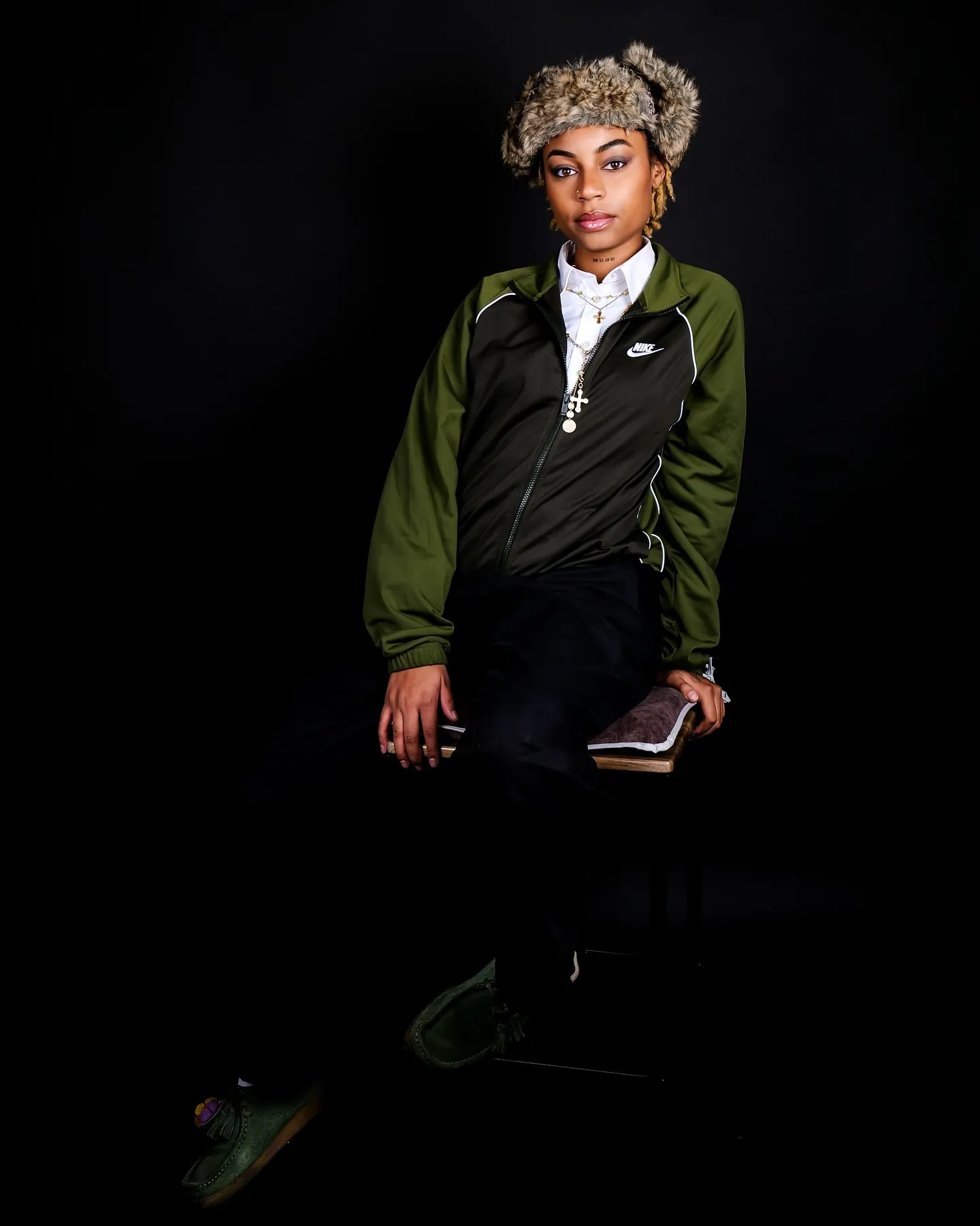 Person in a fur hat and green Nike jacket standing against black, holding a book at their hip