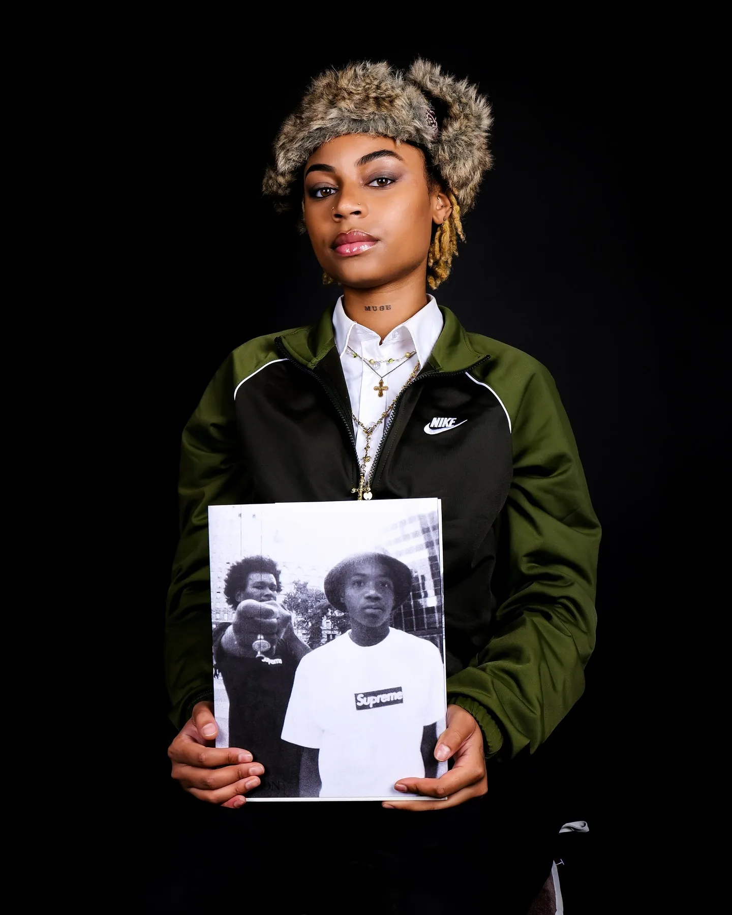 Person in a fur hat and green Nike jacket holding up a black and white photo book with a Supreme shirt