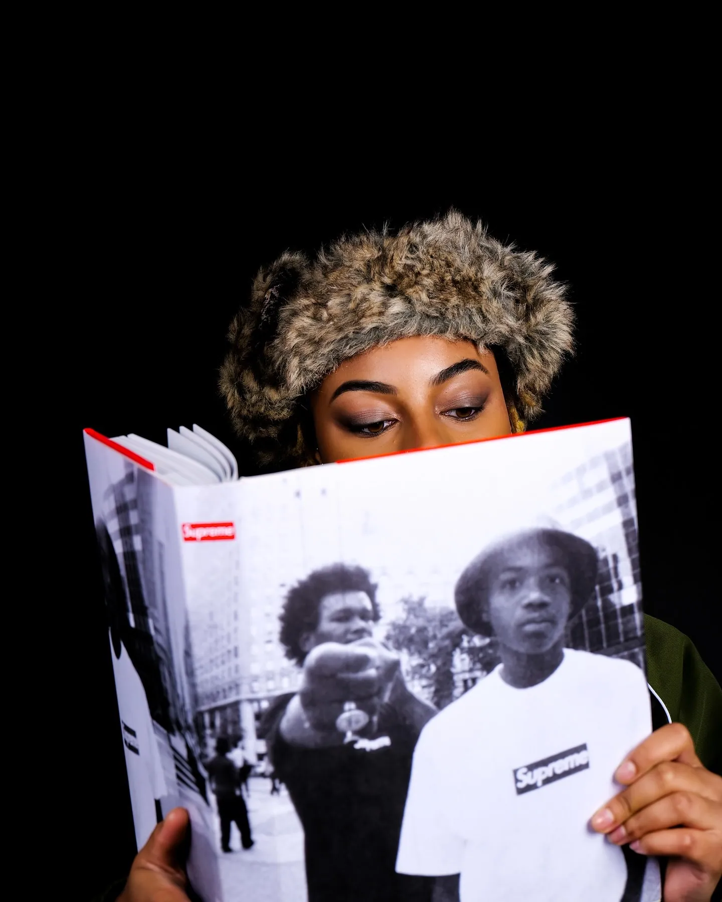 Person in a fur hat peeking over an open Supreme photo book, black background