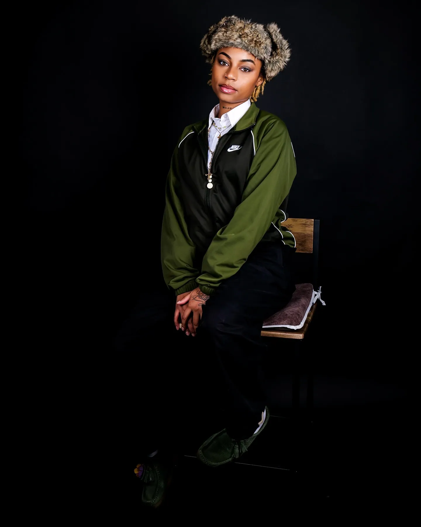 Person in a fur hat and green Nike jacket seated on a wooden chair against a black backdrop