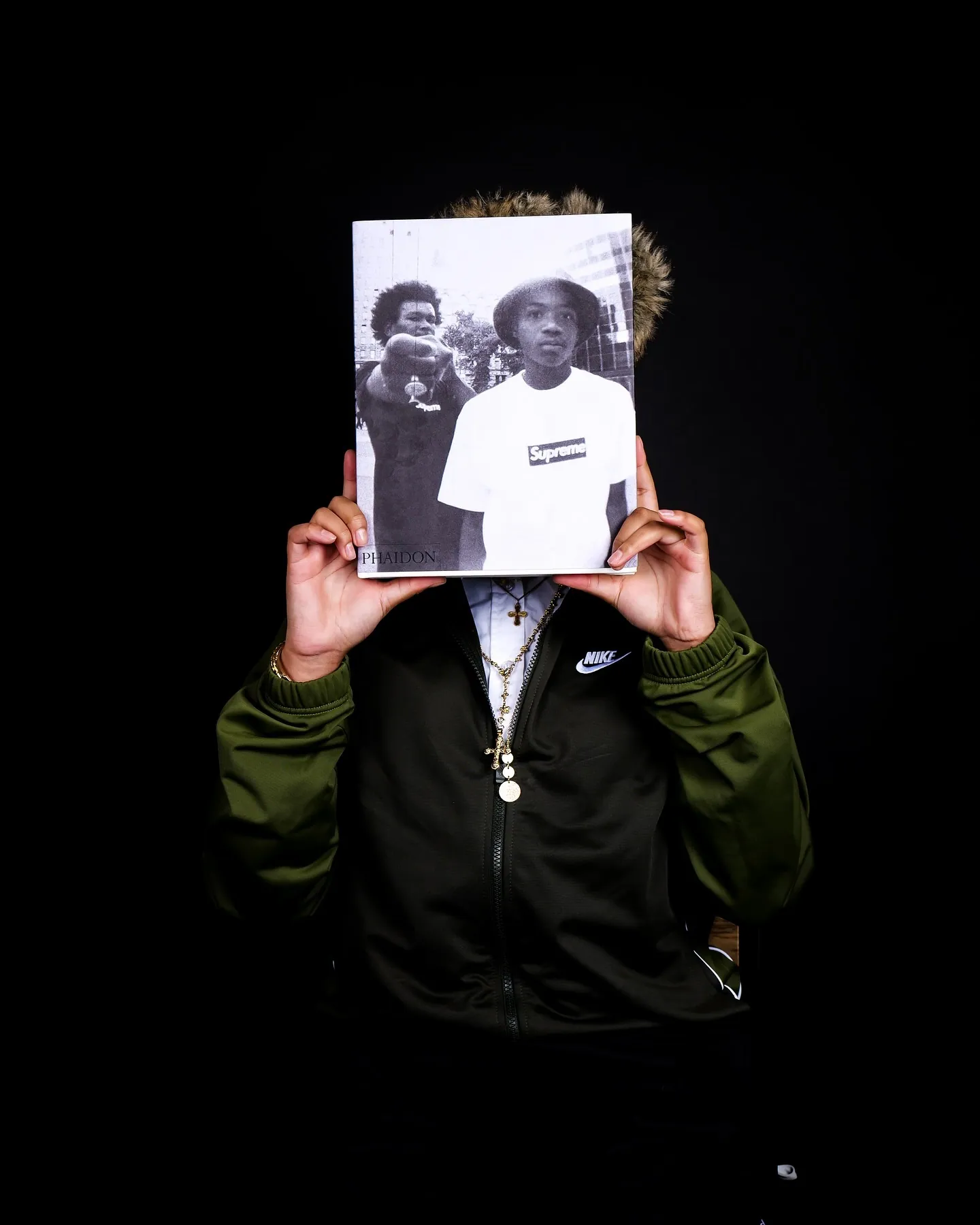 Person holding a black and white Supreme photo book in front of their face, green jacket, black background