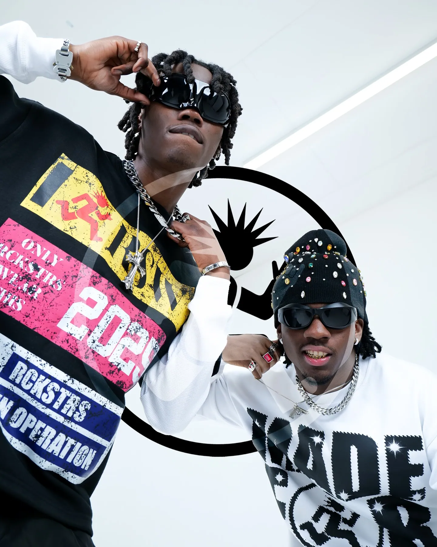 Two men in sunglasses and chains posing close together in graphic RCKSTR tops, a black circle logo behind them