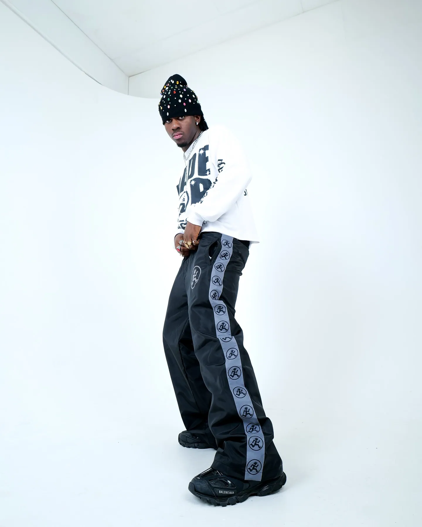 A man in a studded beanie, white graphic top and black logo-striped track trousers posing on a white cyclorama