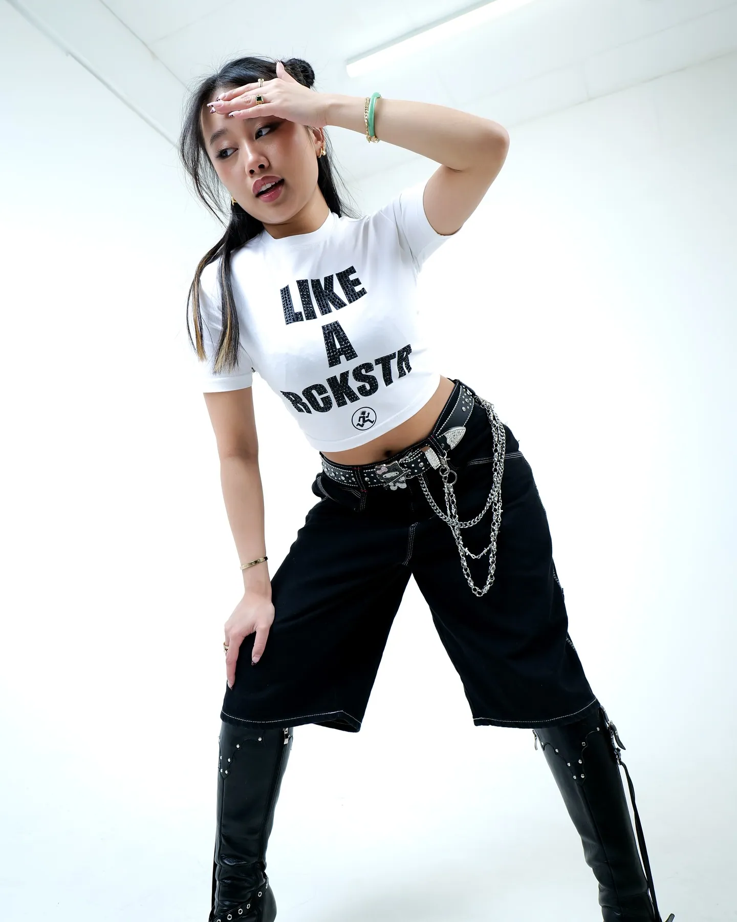A woman in a white LIKE A RCKSTR crop top, black shorts, chain belt and black boots, hand raised to her forehead