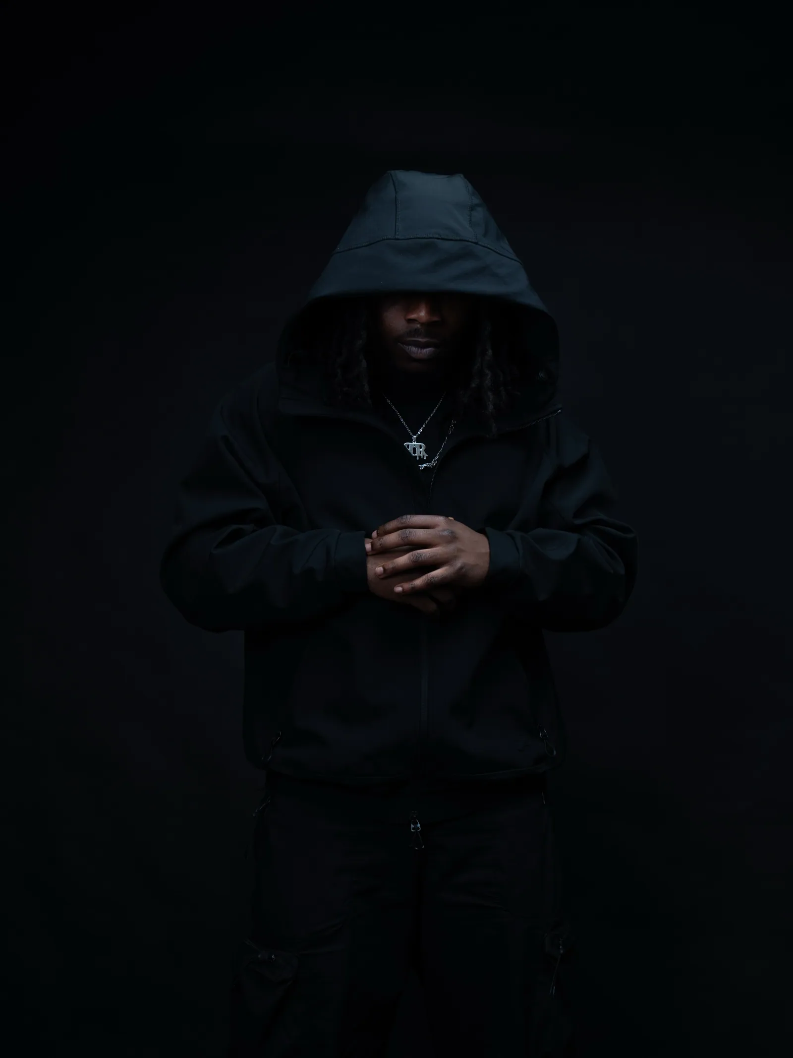 Man in a black hood with hands clasped in front, standing in dim light against a black backdrop