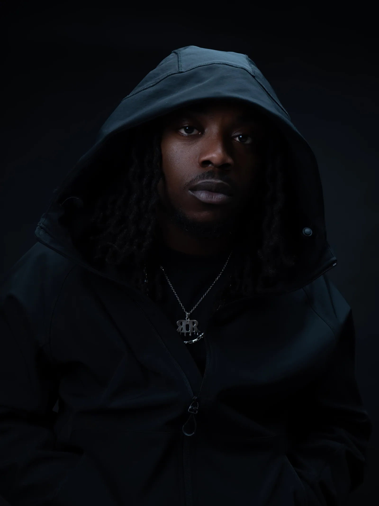 Close portrait of a man in a black hood with a pendant chain, looking at the camera in low light