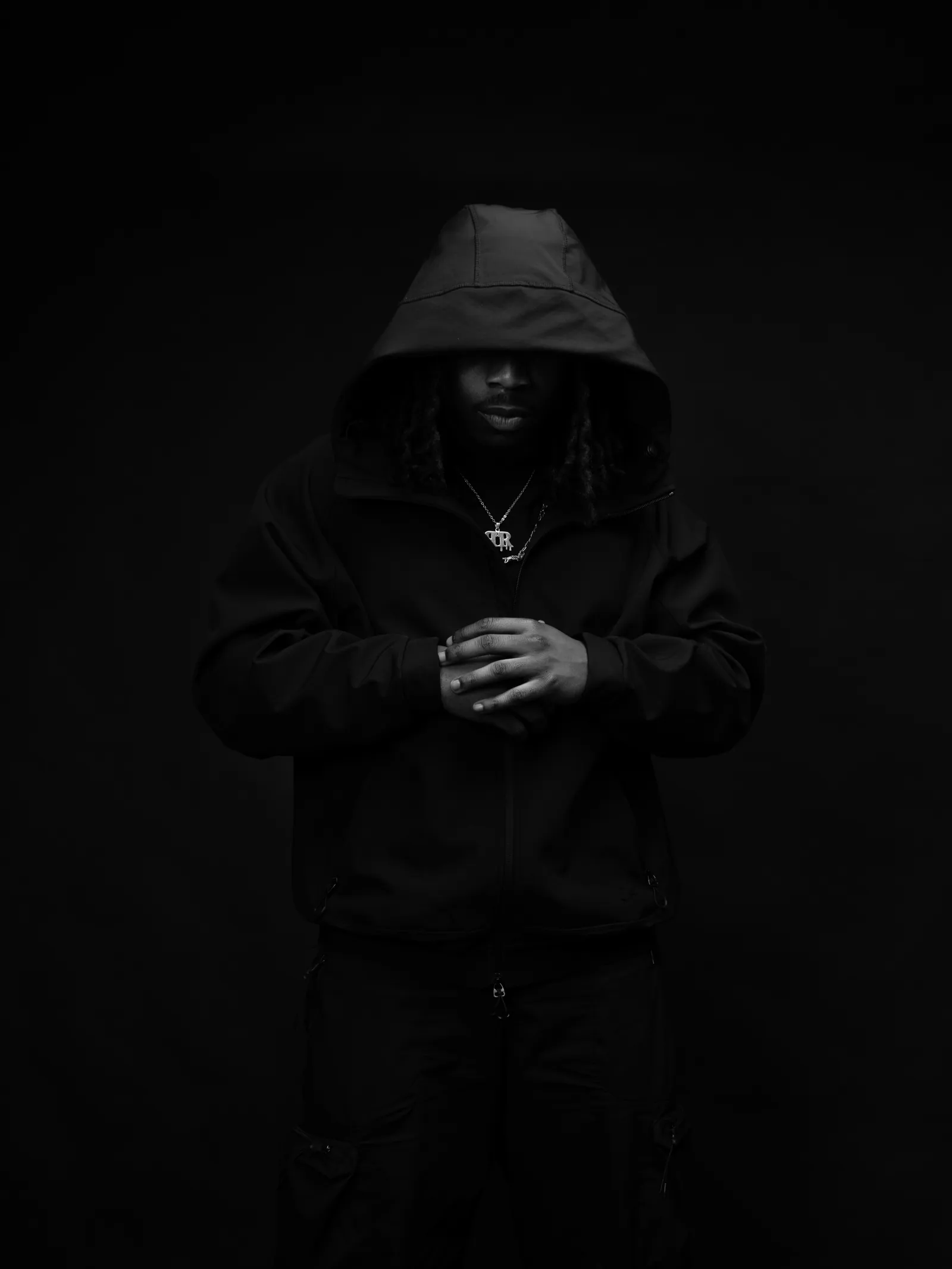 Black and white shot of a hooded man with hands clasped, standing against a dark backdrop