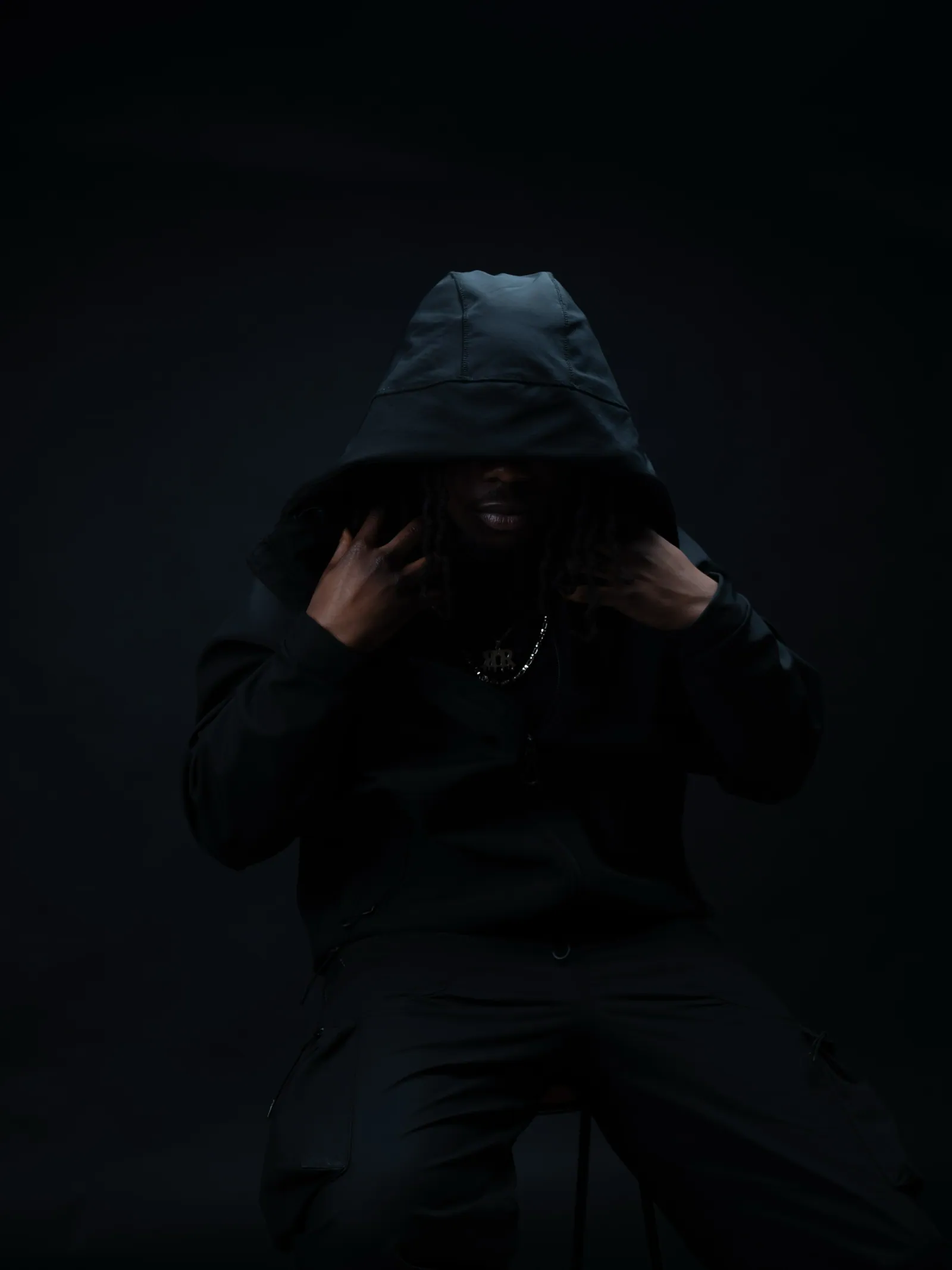 Hooded man seated in the dark with both hands on his hood, face hidden in shadow