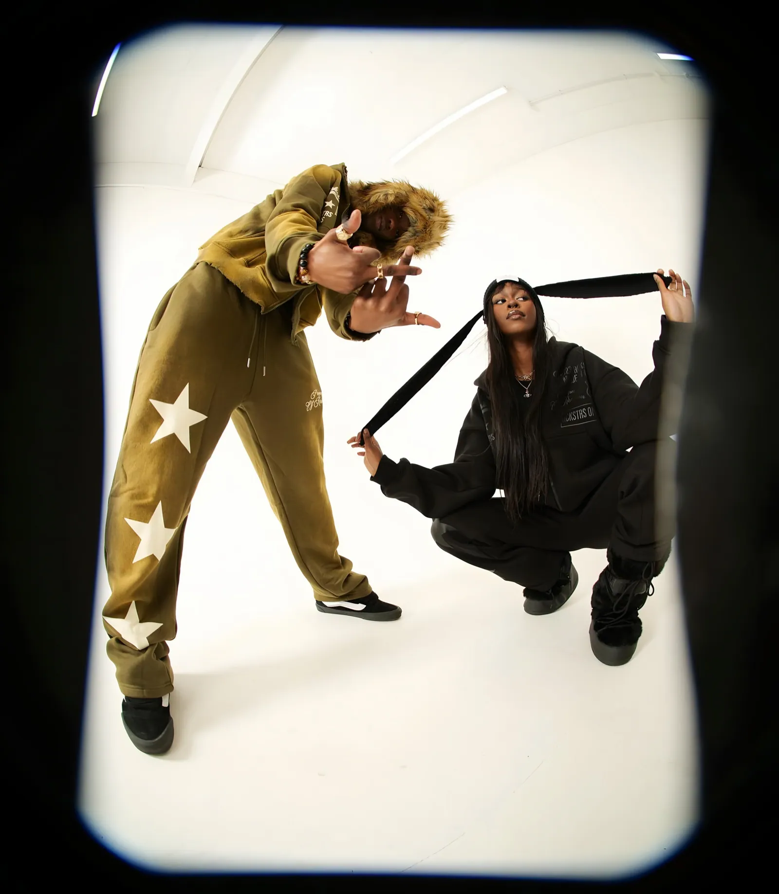 Fisheye view of a man in an olive star-print tracksuit and a woman crouching in black, on a white set