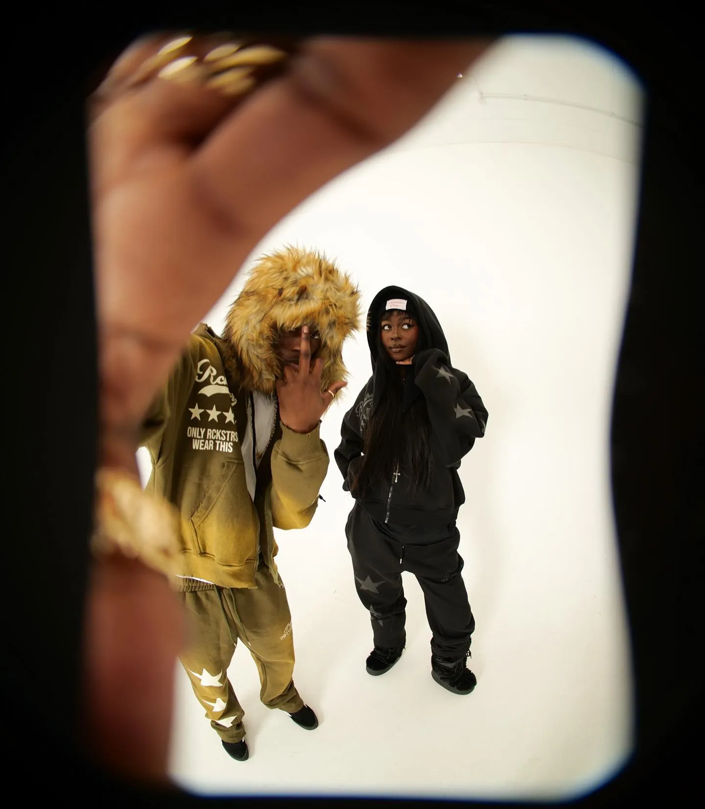 Fisheye shot of a man in a fur-trimmed olive tracksuit and a woman in a black hooded set, on white