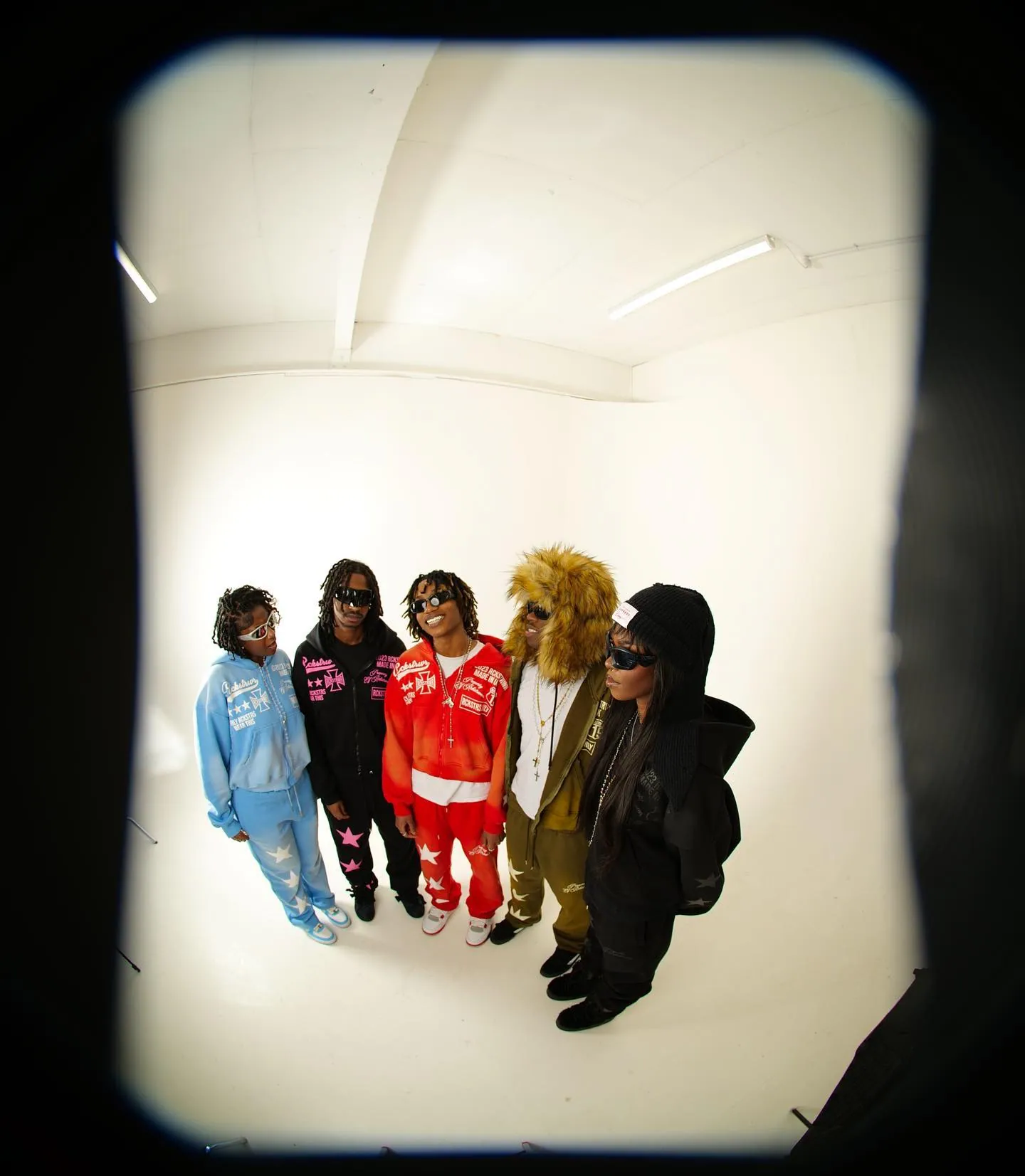 Five people in colourful tracksuits and sunglasses, one in a lion-mane hood, in a white studio, fisheye view