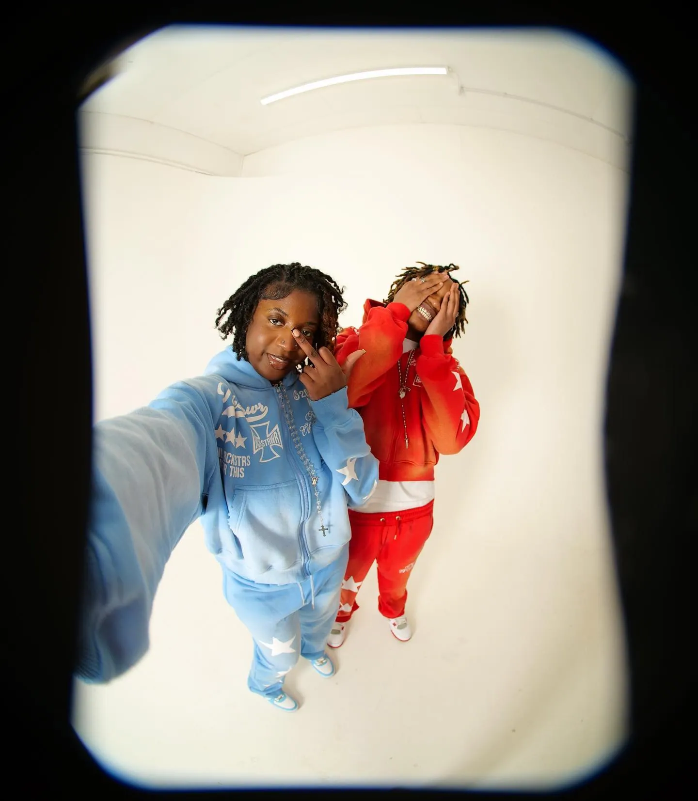 Fisheye shot of two people in matching blue and red star-print tracksuits, hands near their faces, on white
