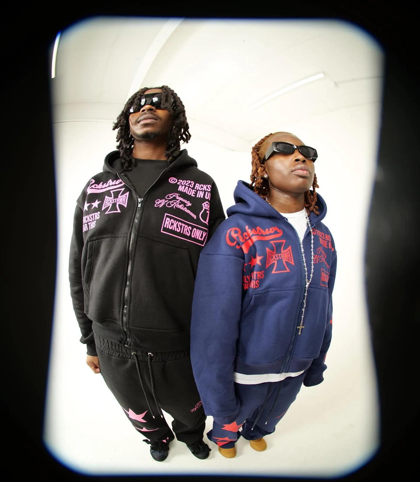 Two people in black and navy tracksuits printed with pink graphics and sunglasses, in a fisheye frame