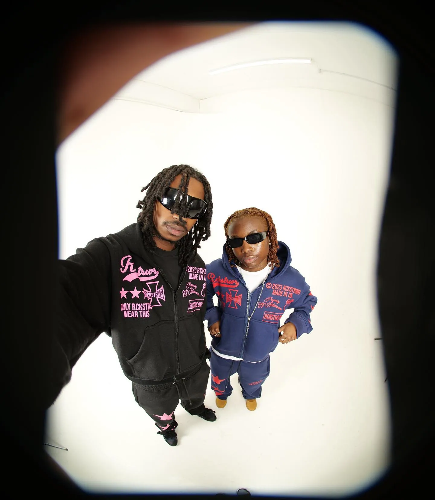 Fisheye shot of a man in a black hoodie with pink print and a person in navy, both in sunglasses, white backdrop