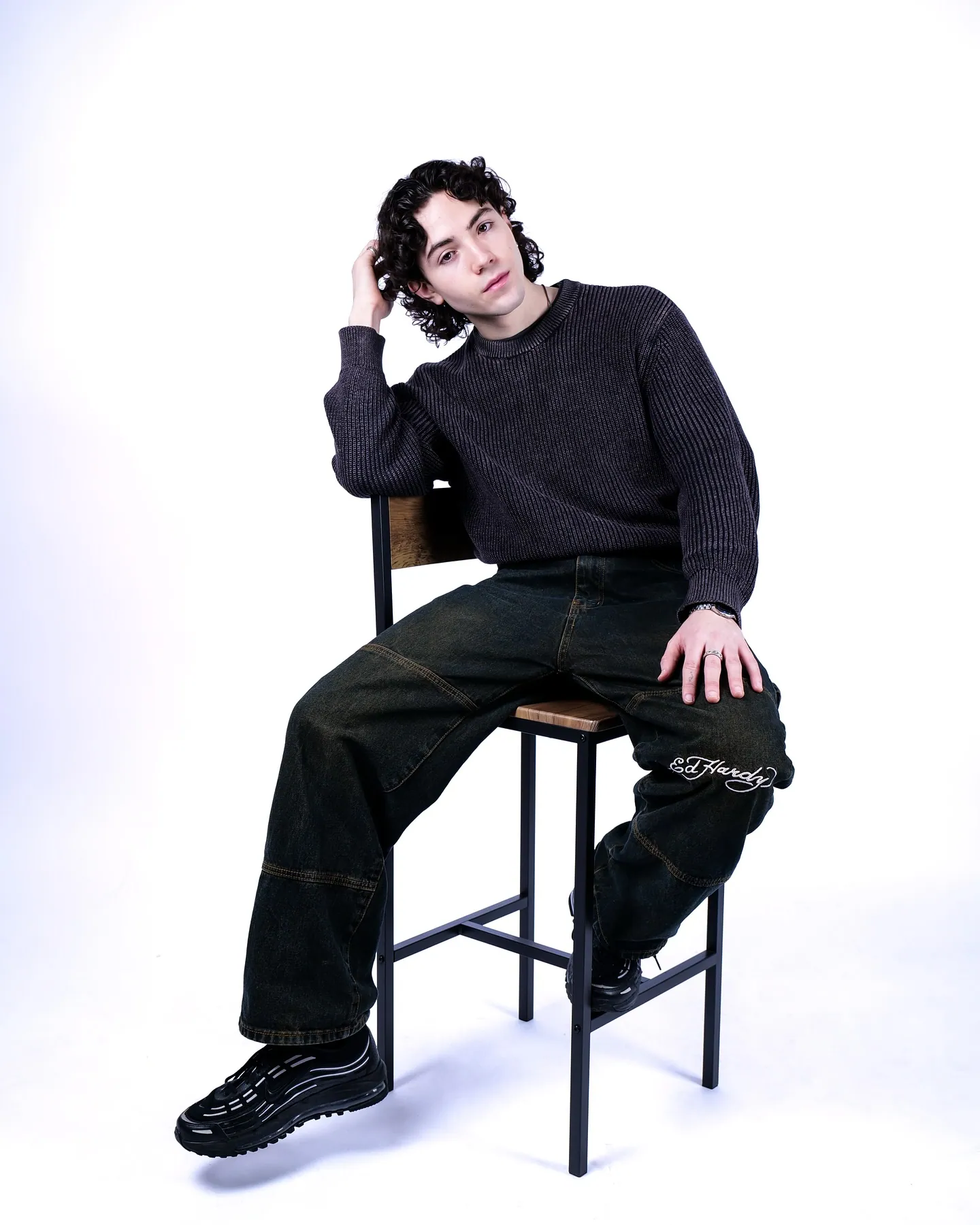 A young man with curly hair on a high stool in a dark knit jumper and baggy dark jeans, hand in his hair, pale backdrop