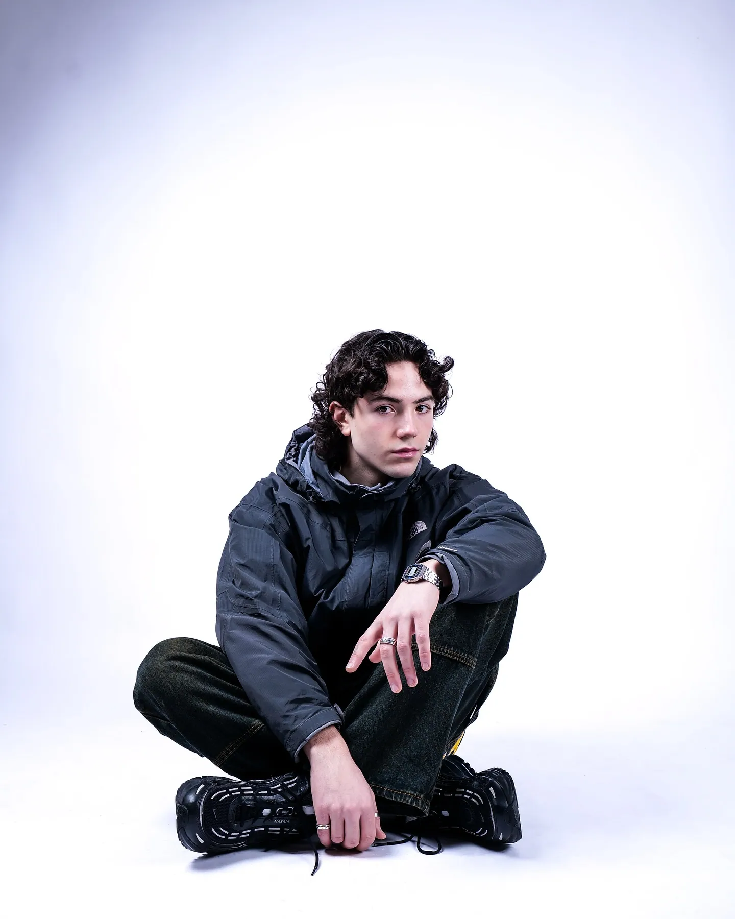 A young man with curly hair sitting cross-legged on the floor in a dark jacket and trainers, hand drooping over his knee
