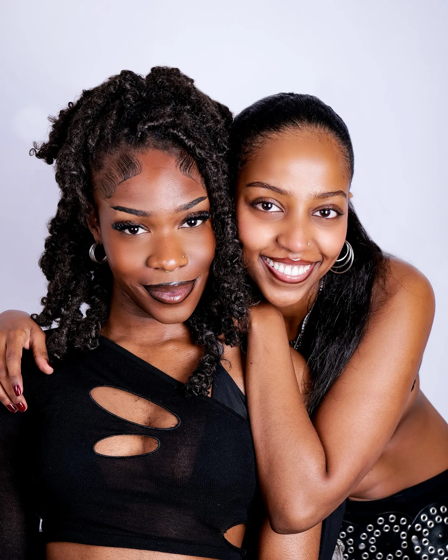 Two smiling women posing cheek to cheek in black outfits and hoop earrings against a lilac backdrop