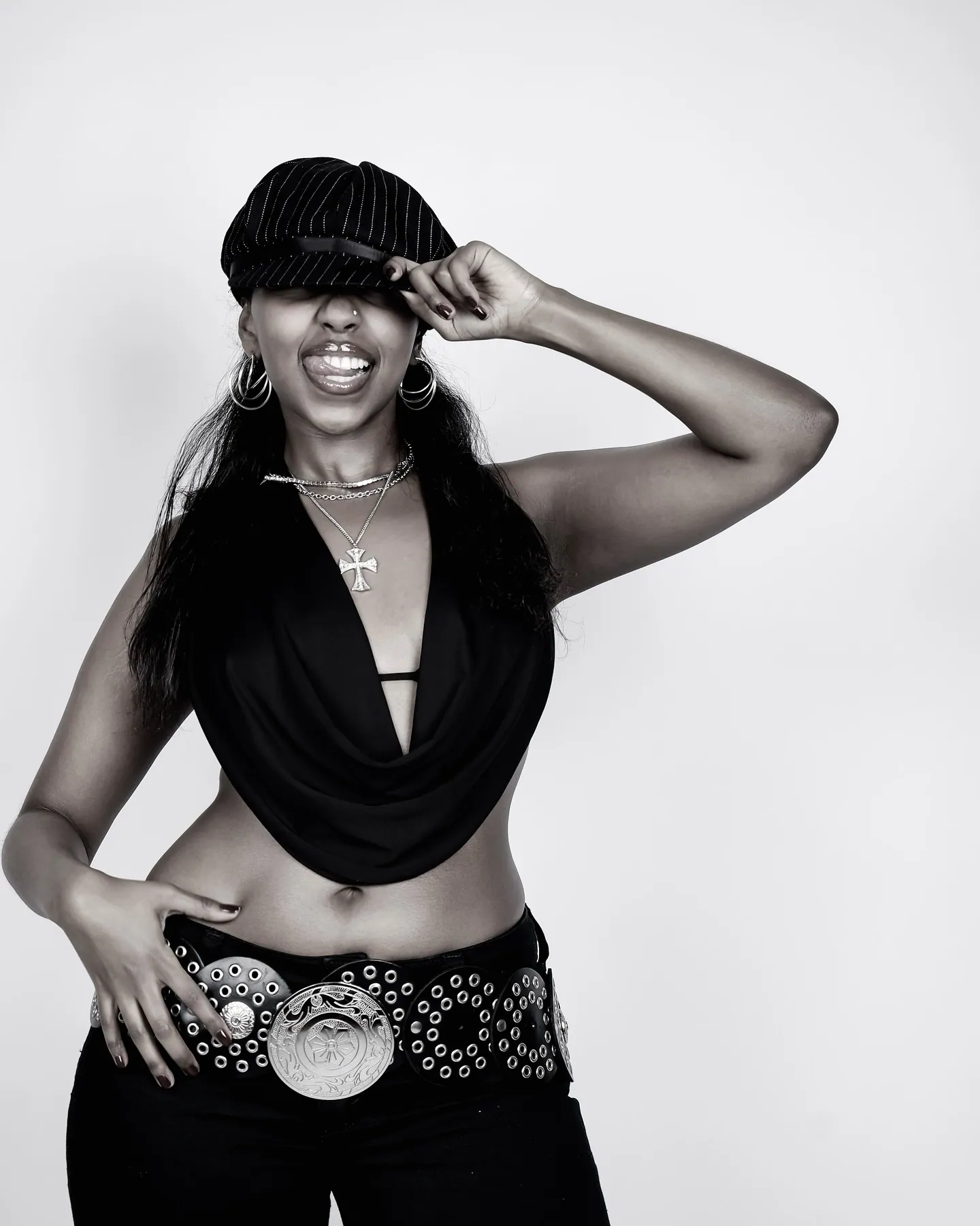 Black and white portrait of a woman in a pinstripe cap and cowl top, a hand on her hip, studio backdrop