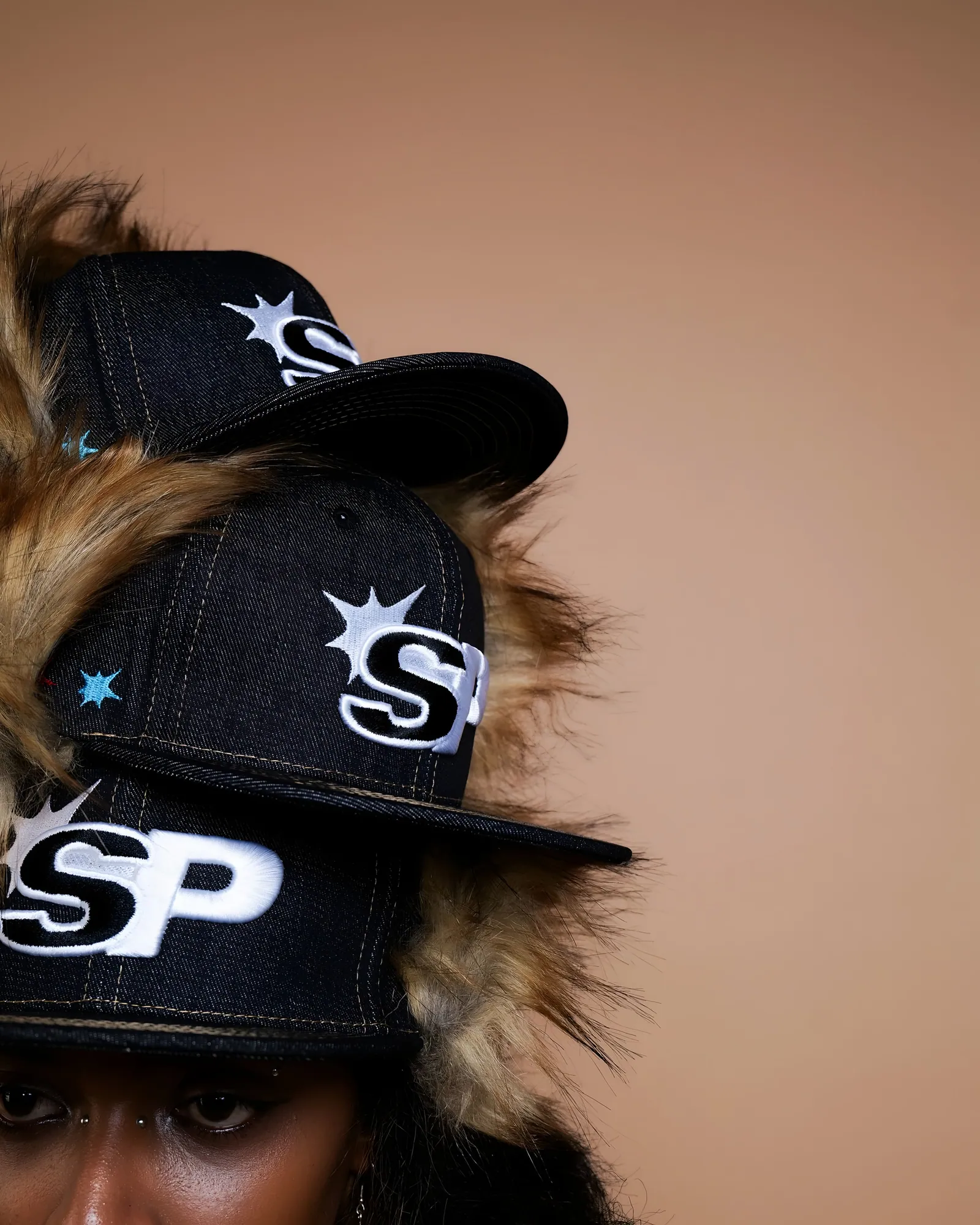 Three black denim caps with SP star logos stacked over a faux fur hood, a face cropped at the bottom, beige backdrop