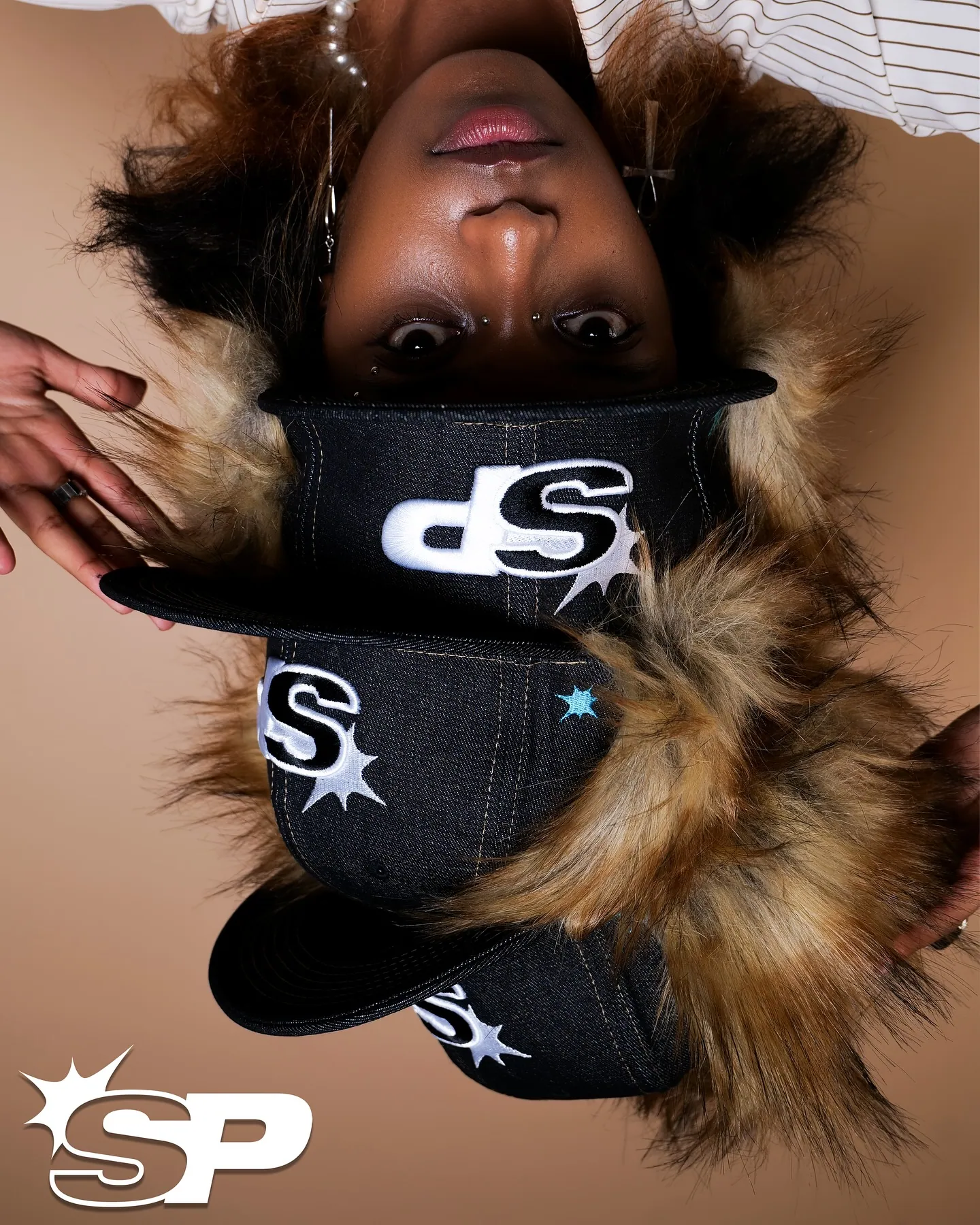 Upside-down portrait of a person wearing stacked SP denim caps with a fur trim, SP logo in the corner, beige backdrop