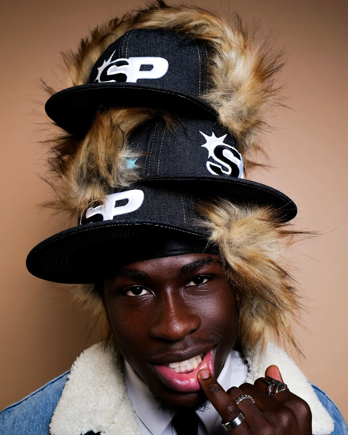 A man in a denim sherpa jacket and tie wearing three stacked SP caps with faux fur, touching his lip, beige backdrop