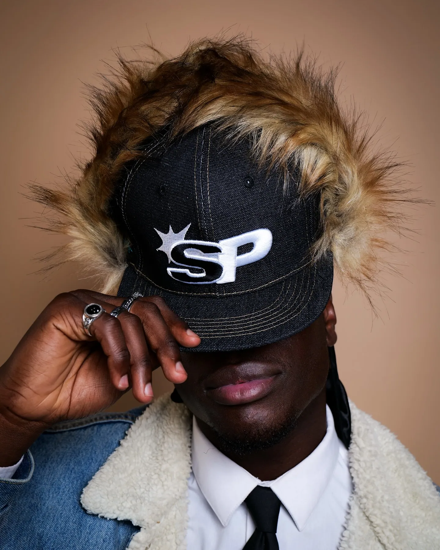 A man in a white shirt, tie and denim sherpa jacket pulling an SP cap with fur trim over his eyes, beige backdrop