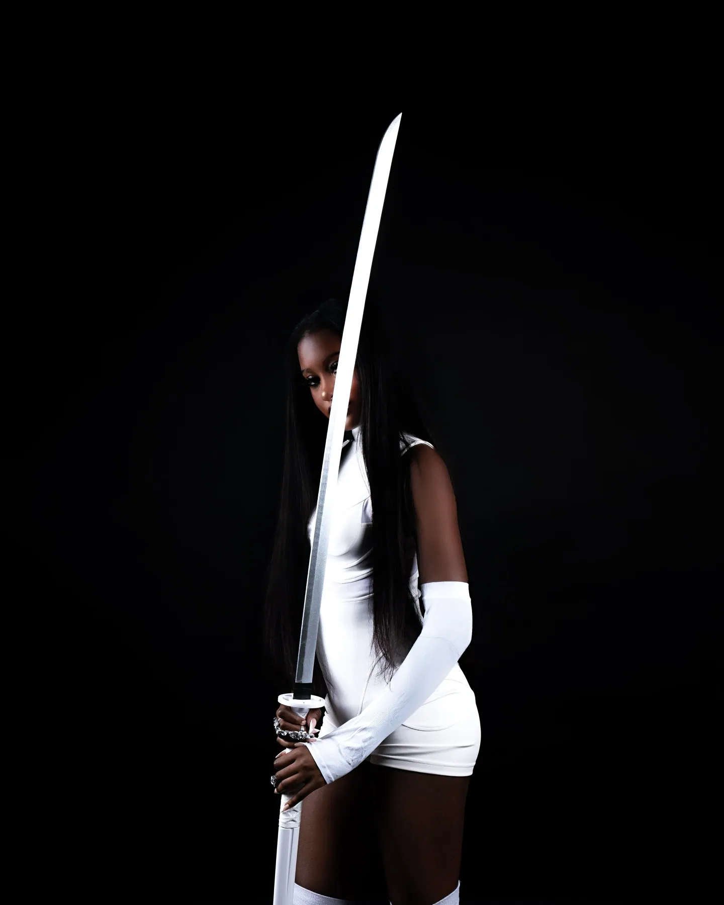 A woman in a white fitted dress and white glove holding a katana upright in front of her face against a black backdrop