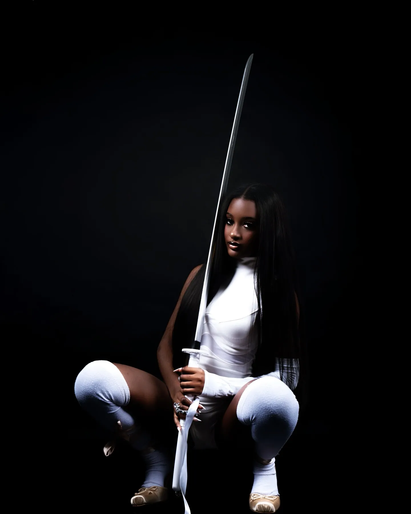 A woman in white crouching in thigh-high socks with a katana held upright, long dark hair, black backdrop