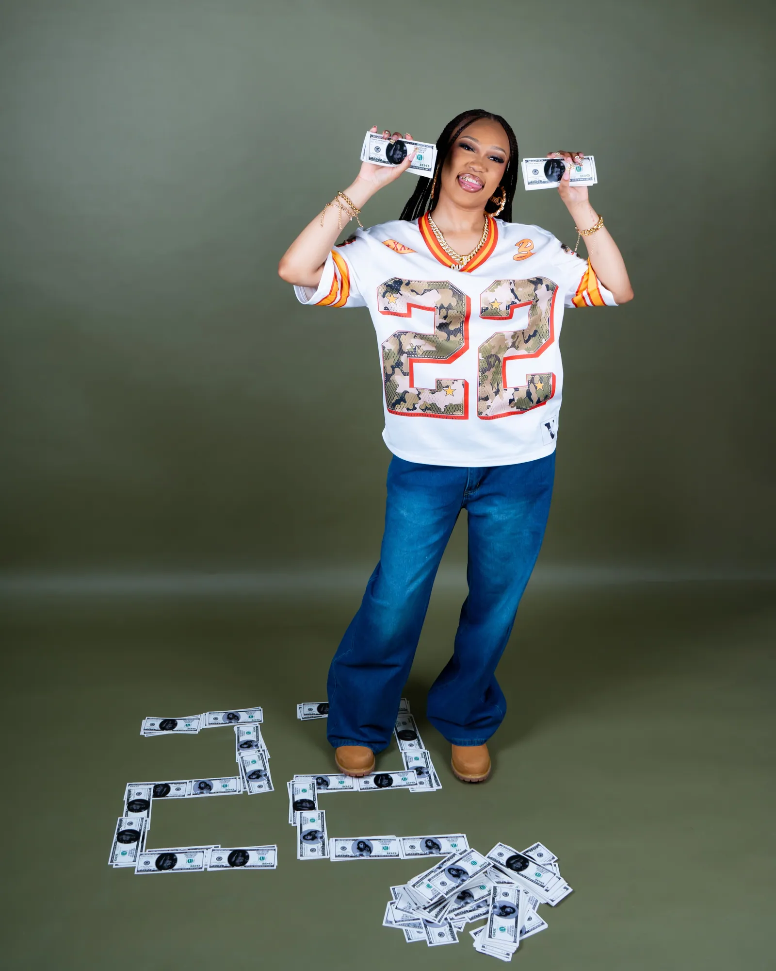 A woman in a number 22 jersey and jeans holding two bundles of cash, notes spelling 22 on the floor, green backdrop