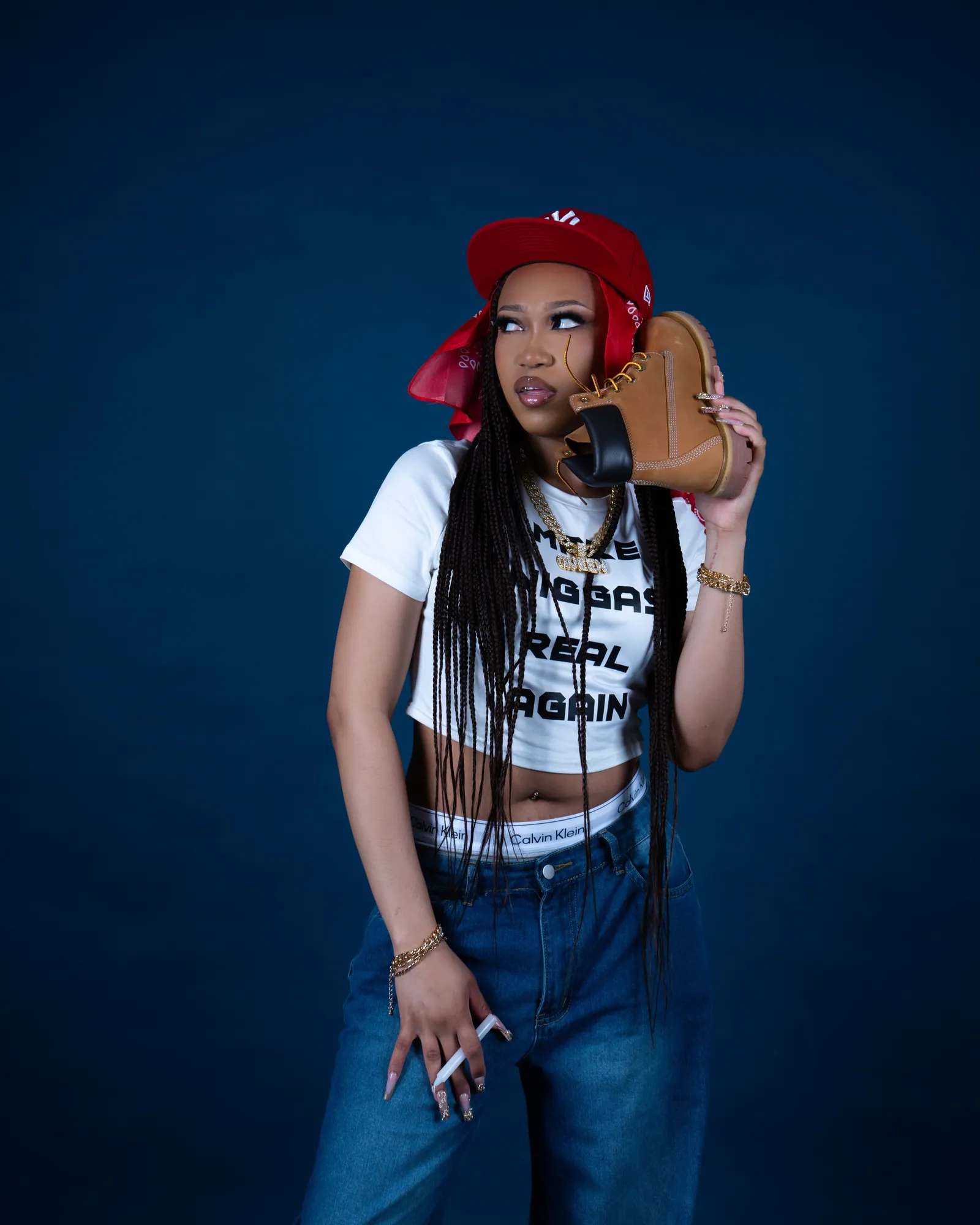 A woman in a red cap and white slogan crop top holding a tan boot to her ear like a phone, dark blue backdrop