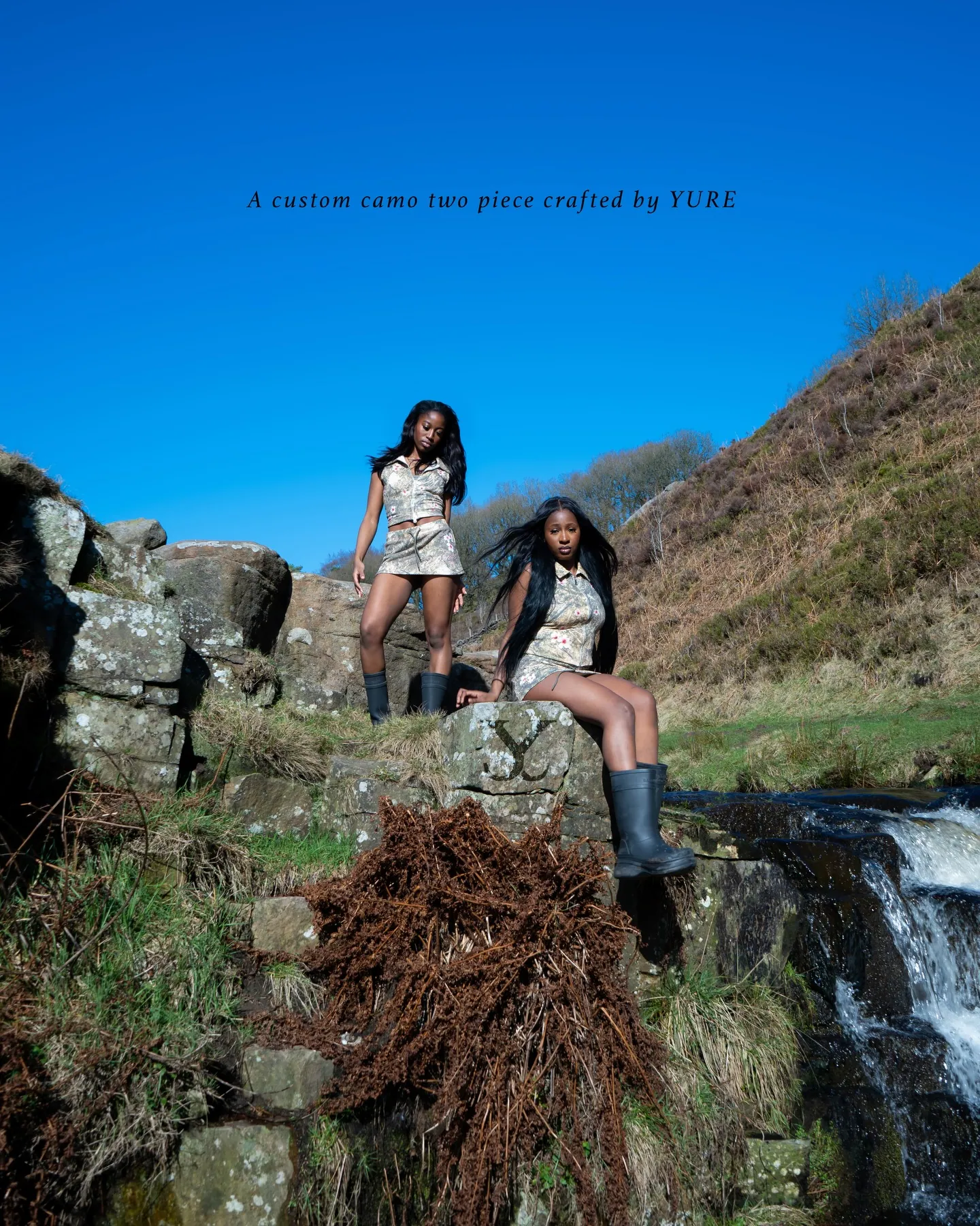 Two models in camo two-piece outfits and wellies on rocks by a stream, with text A custom camo two piece crafted by YURE