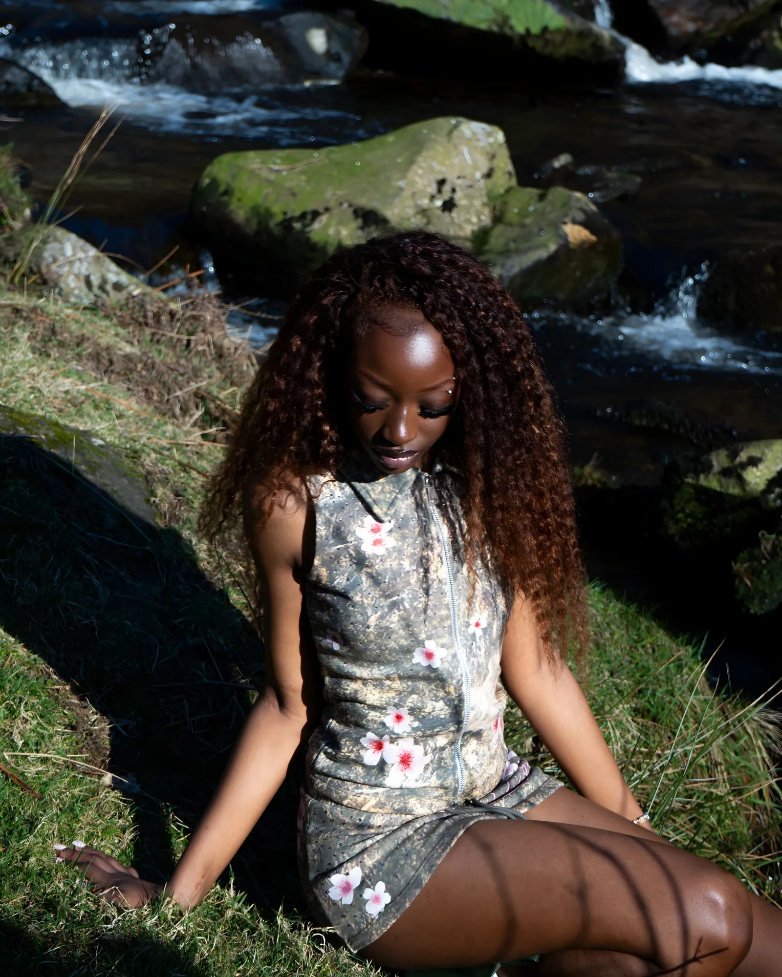 Model with long curly hair in a floral camo outfit sitting on a grassy bank beside a rocky stream in sunlight