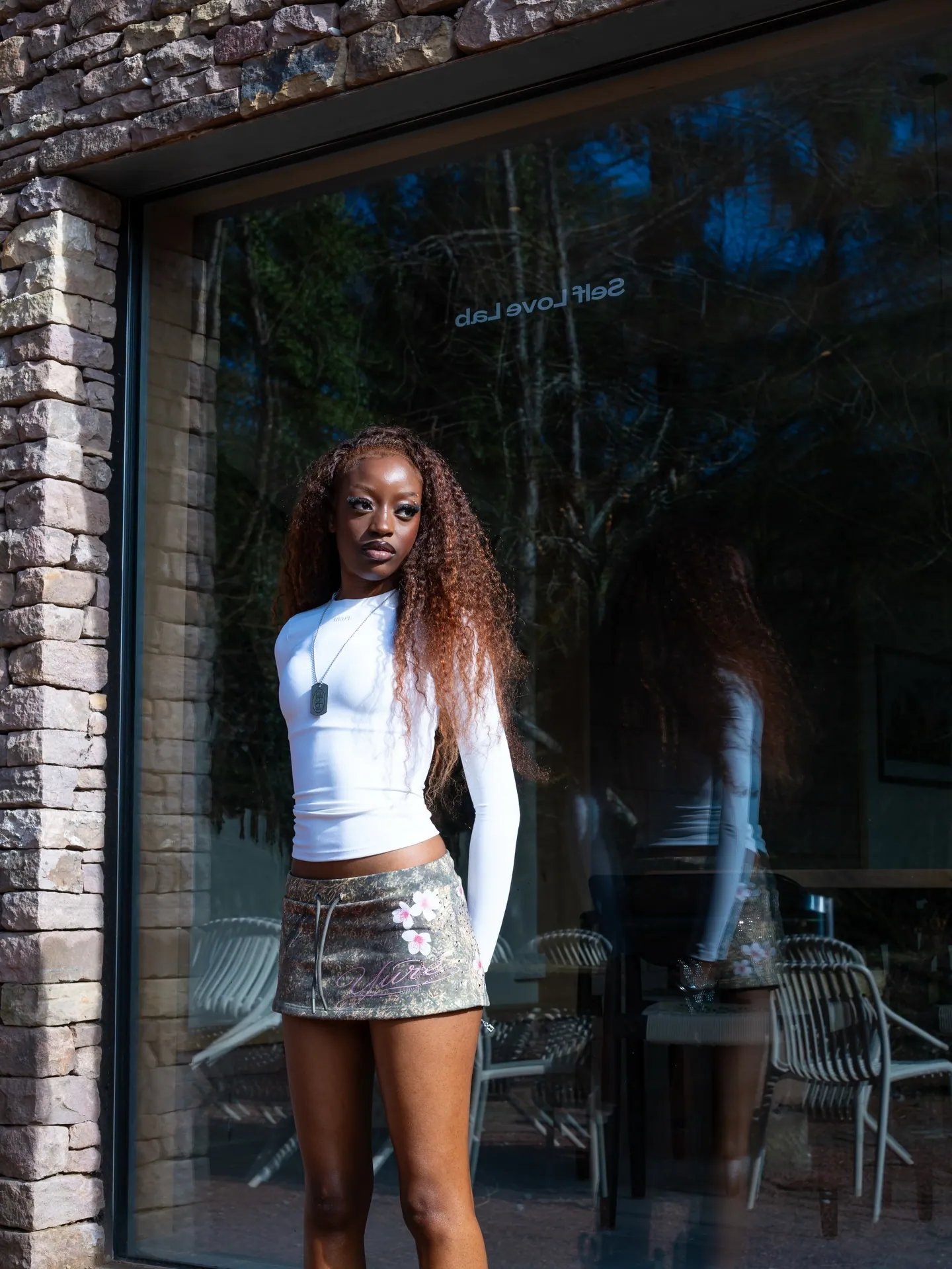 Model with long curly hair in a white long-sleeve top and camo mini skirt in front of a glass wall reading Self Love Lab