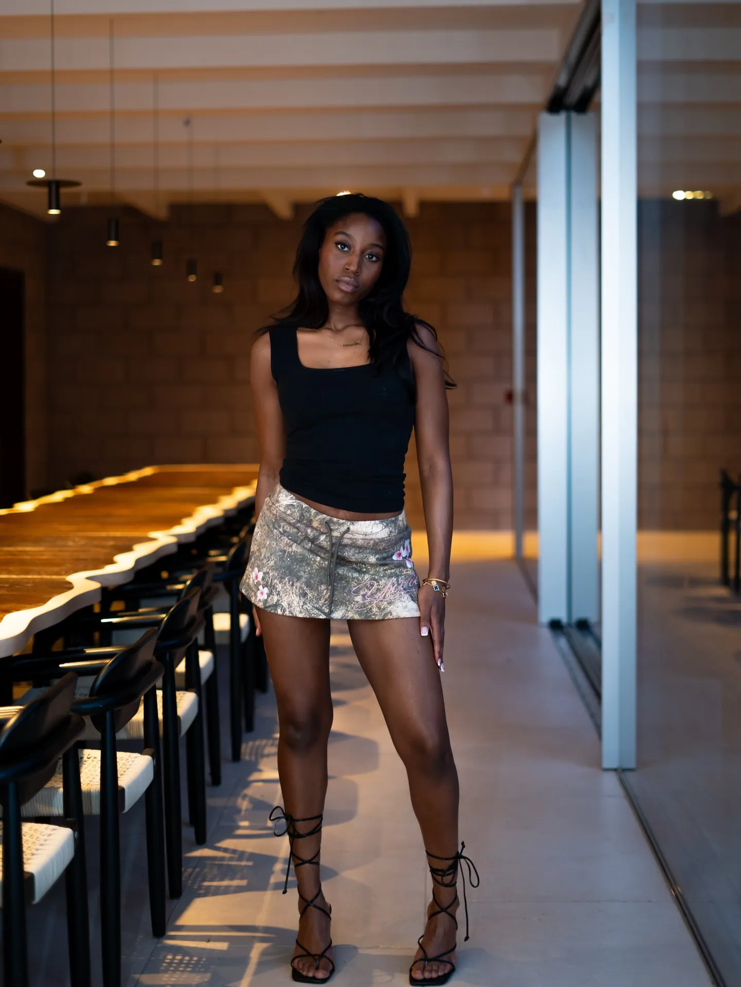 Model in a black vest top, camo mini skirt and black lace-up sandals standing beside a long wooden table and chairs