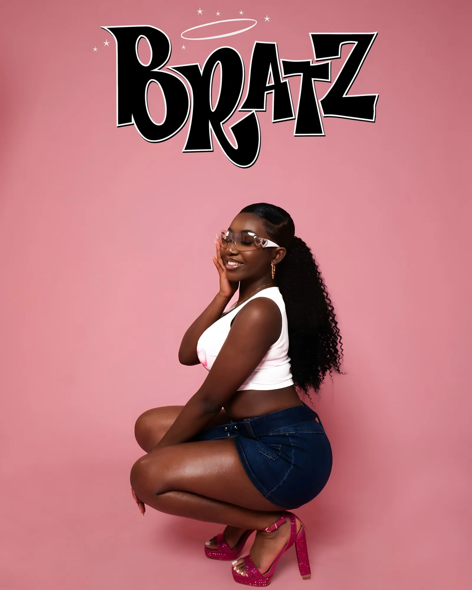 Woman crouching in front of a pink backdrop under BRATZ lettering, in a white top, denim skirt and pink heels