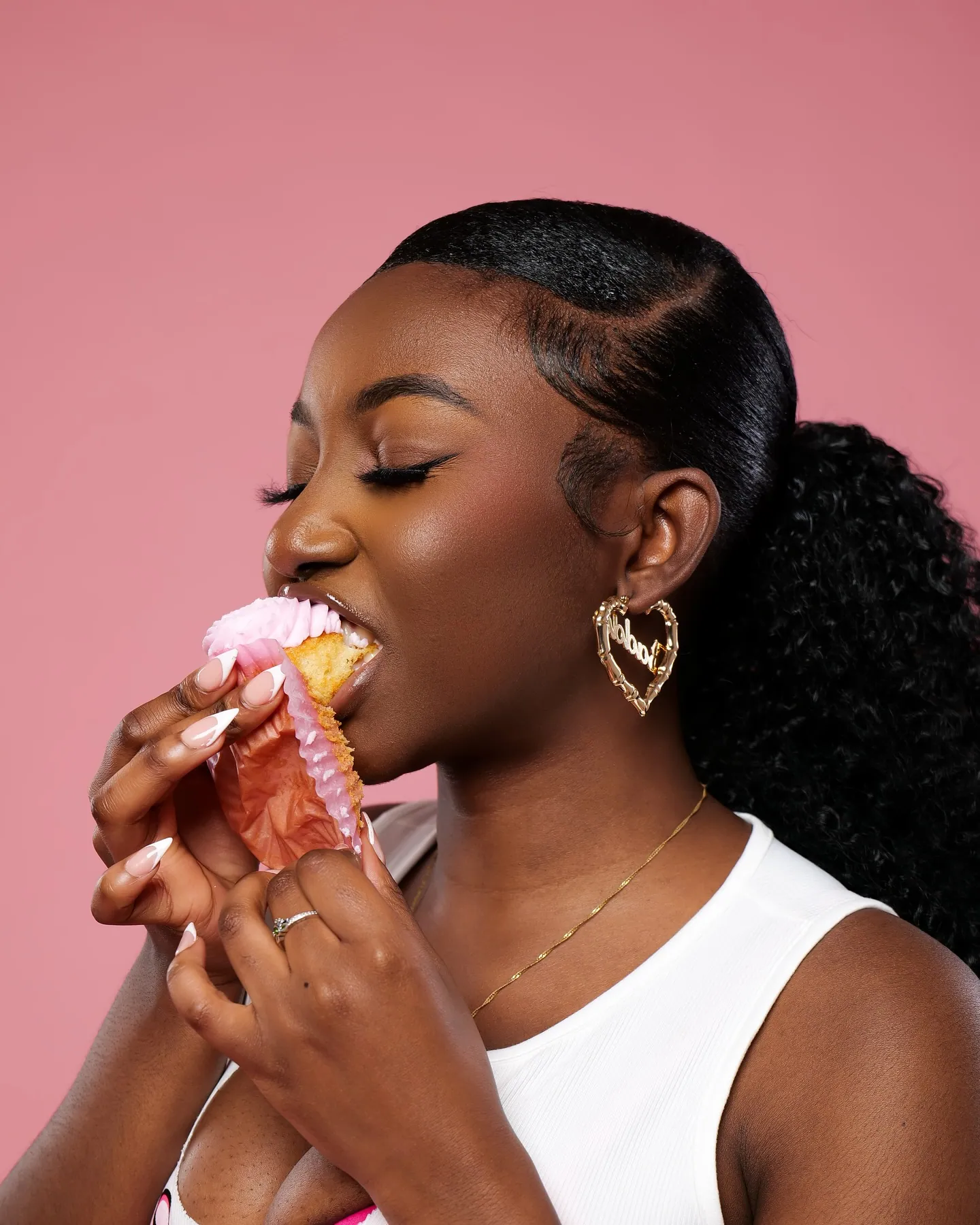 Close profile of a woman with a high curly ponytail and heart earrings biting into a cupcake, pink backdrop