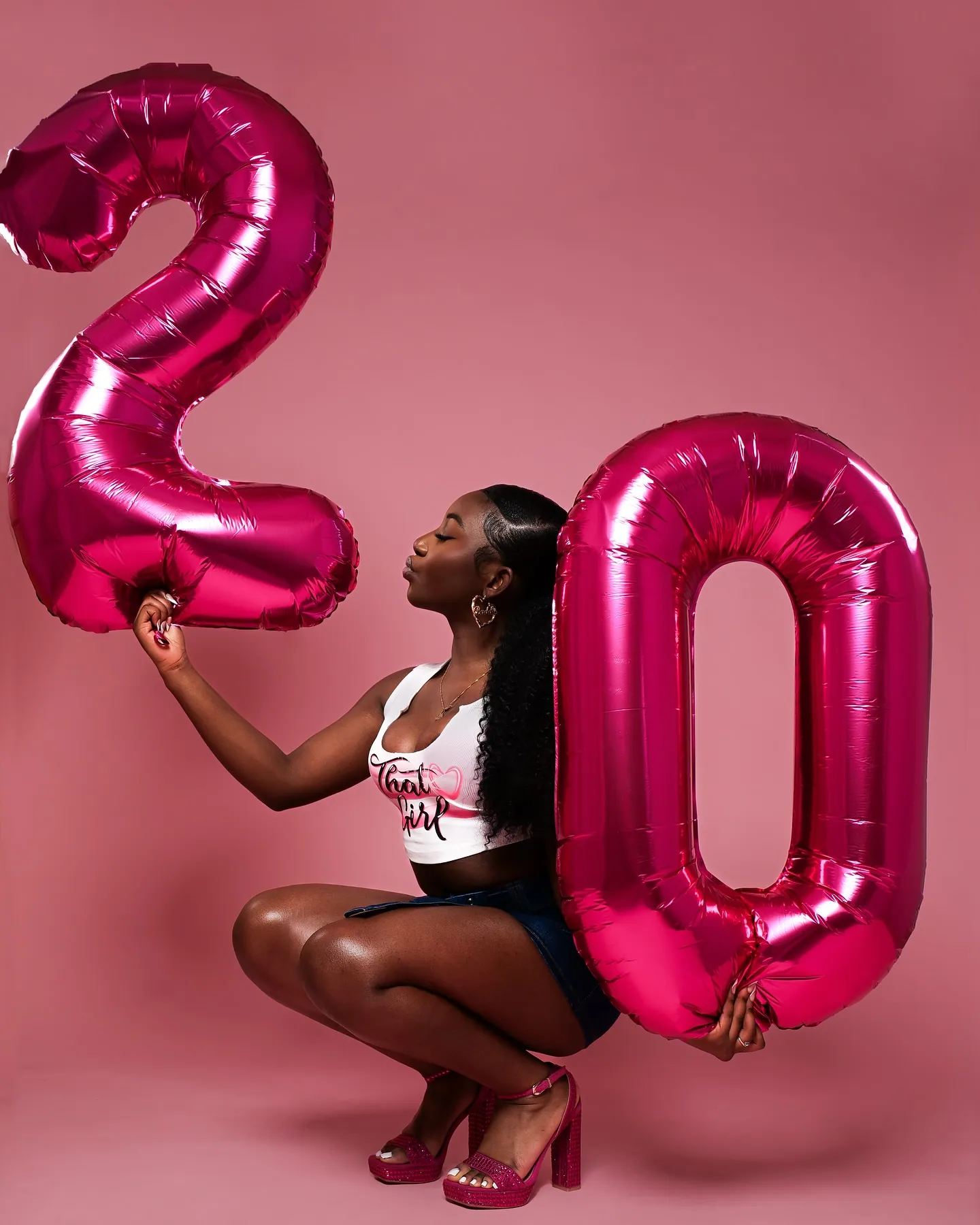 Woman crouching between two pink foil balloons that make the number 20, pink studio backdrop