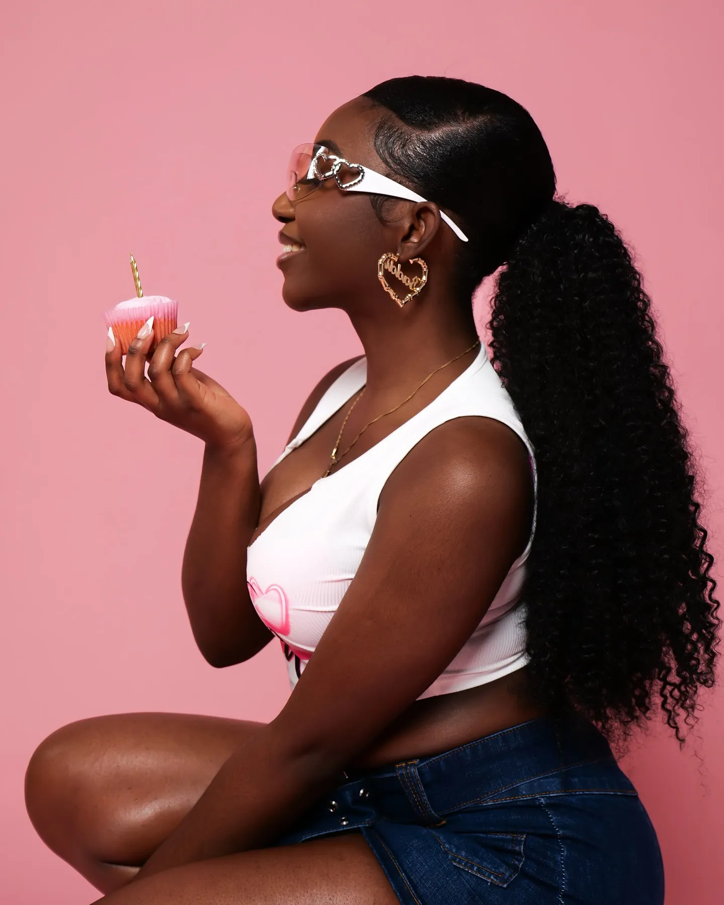 Profile of a woman with heart-framed sunglasses and a curly ponytail holding a cupcake with a candle