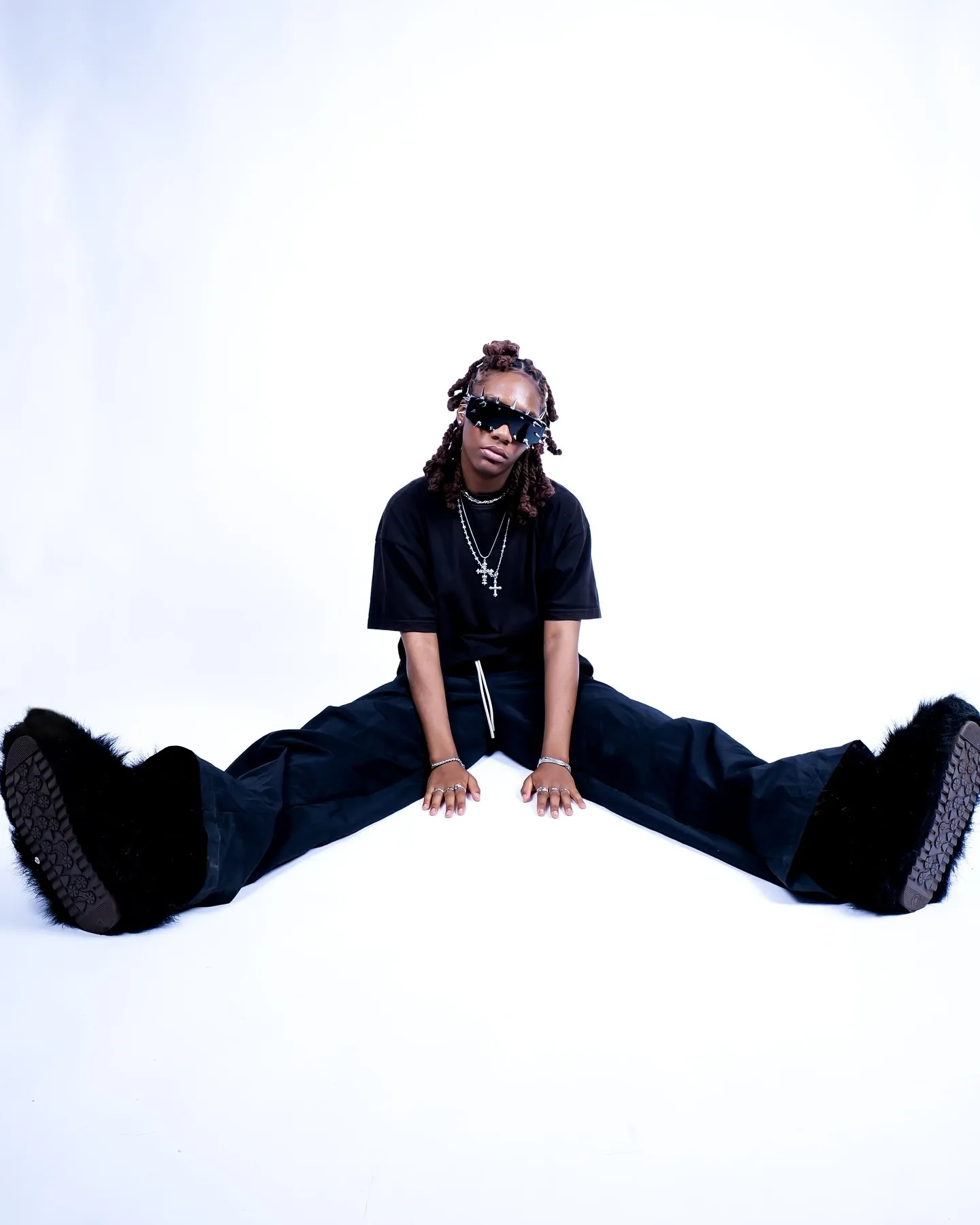 A model sitting on the floor with legs wide apart in black clothes, sunglasses and fluffy black boots, white backdrop